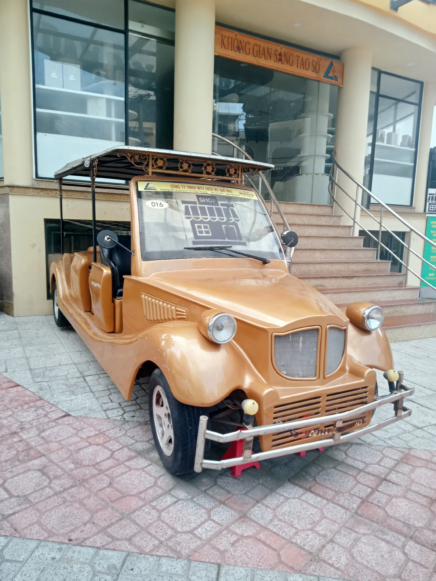 A low-angle, medium shot reveals a gold-colored, vintage-style electric car parked on a stone-tiled surface. The car is positioned in front of a building with a modern facade, featuring large glass windows and tan-colored pillars. A flight of stairs with a metal railing leads to the building's entrance. Above the entrance, a sign reads "KHÔNG GIAN SÁNG TẠO SỐ 24". The car displays stickers and numbers on its windshield, with the text "CÔNG TY TNHH MTV DỊCH VỤ SAO MAI" and the number "016" visible. In the background, the reflection in the glass windows suggests an interior with shelves and other objects. The scene appears to be in the Nha Trang Ward area of Vietnam.