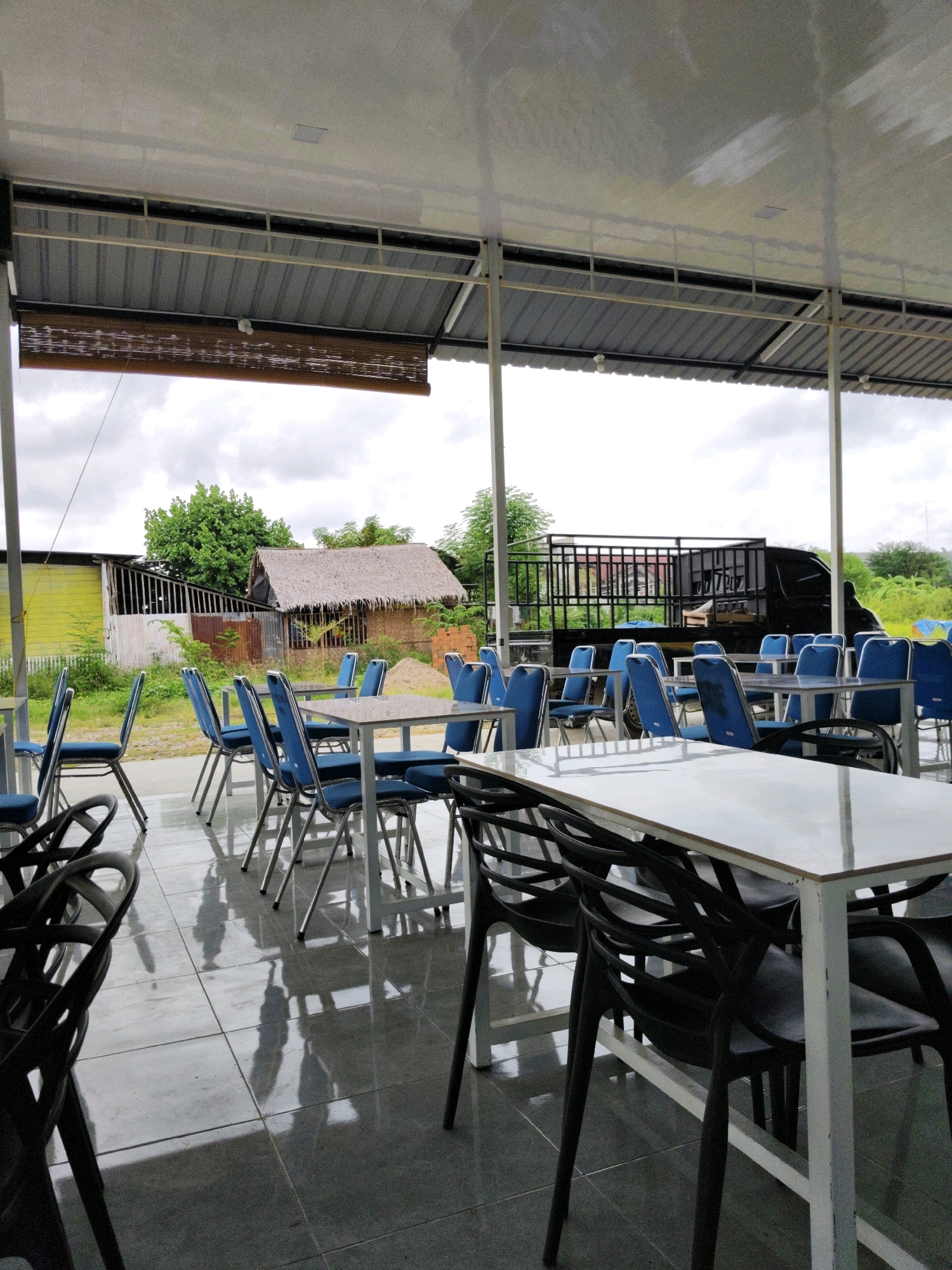 The image depicts an outdoor seating area, likely part of a restaurant or cafe, in Lhokseumawe, Indonesia. The area is covered by a corrugated metal roof supported by white metal posts. A bamboo blind hangs from the edge of the roof, providing shade. The floor is tiled and reflective, showing shadows of the tables and chairs.
Numerous tables with white tops and metal legs are arranged throughout the space, each accompanied by blue upholstered chairs with metal frames. Some tables are empty, while others appear to have been recently used, with a few chairs slightly out of place.
In the background, a black pickup truck is parked. Beyond the seating area, there are buildings, including a structure with a thatched roof and another with a yellow-painted wall. Lush green trees and vegetation surround the scene, suggesting a rural or semi-rural setting. The sky is overcast, with a cloudy sky visible. The overall impression is one of a simple, functional outdoor dining establishment.