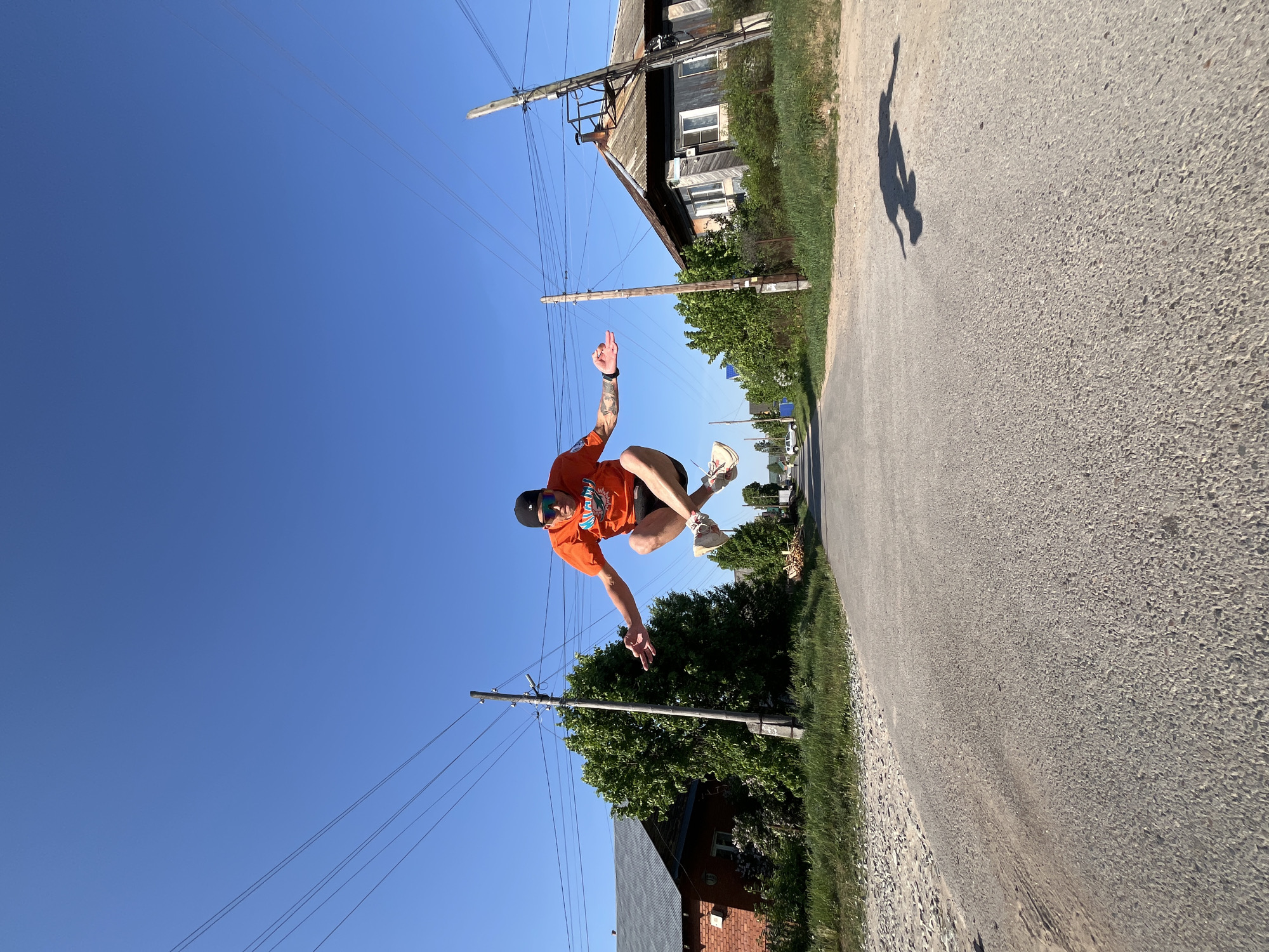 A person is mid-jump in an outdoor environment on a sunny day. The location appears to be a paved road in a rural or suburban setting. The person is wearing an orange shirt, dark shorts, sneakers, and a baseball cap. They have tattoos on their left arm. The person's shadow is visible on the road.
The background includes utility poles with power lines, trees, and houses. The houses are of varying construction, including what appears to be a wooden house with multiple windows and a brick house. The overall impression is of a day in a residential area.
It is not possible to definitively confirm the exact location as Chaikovsky, Russia without additional information or metadata.