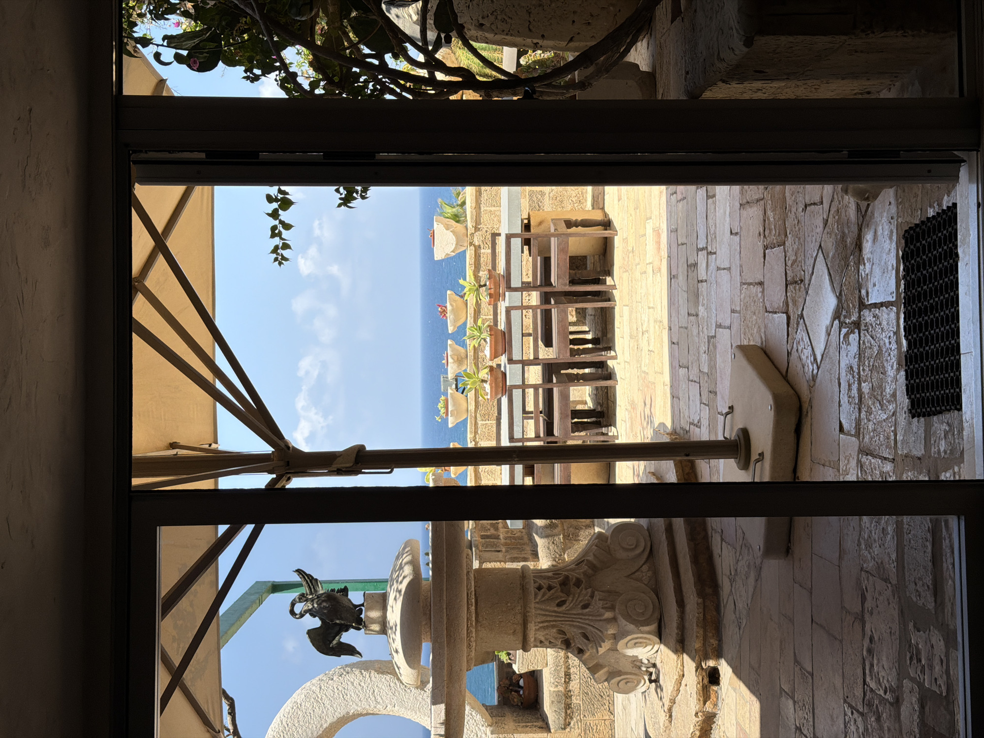 This image depicts an outdoor patio with a view of the Mediterranean Sea, likely located in Tel Aviv, Israel, given the architectural style and the presence of a fountain with a bird sculpture. The patio is paved with stone tiles and features a stone fountain with a bird figurine, a large umbrella providing shade, a dining table with chairs, and several potted plants. The sea is visible in the background, with a clear blue sky above. The scene is captured through an open glass door, suggesting an interior space looking out onto the patio.