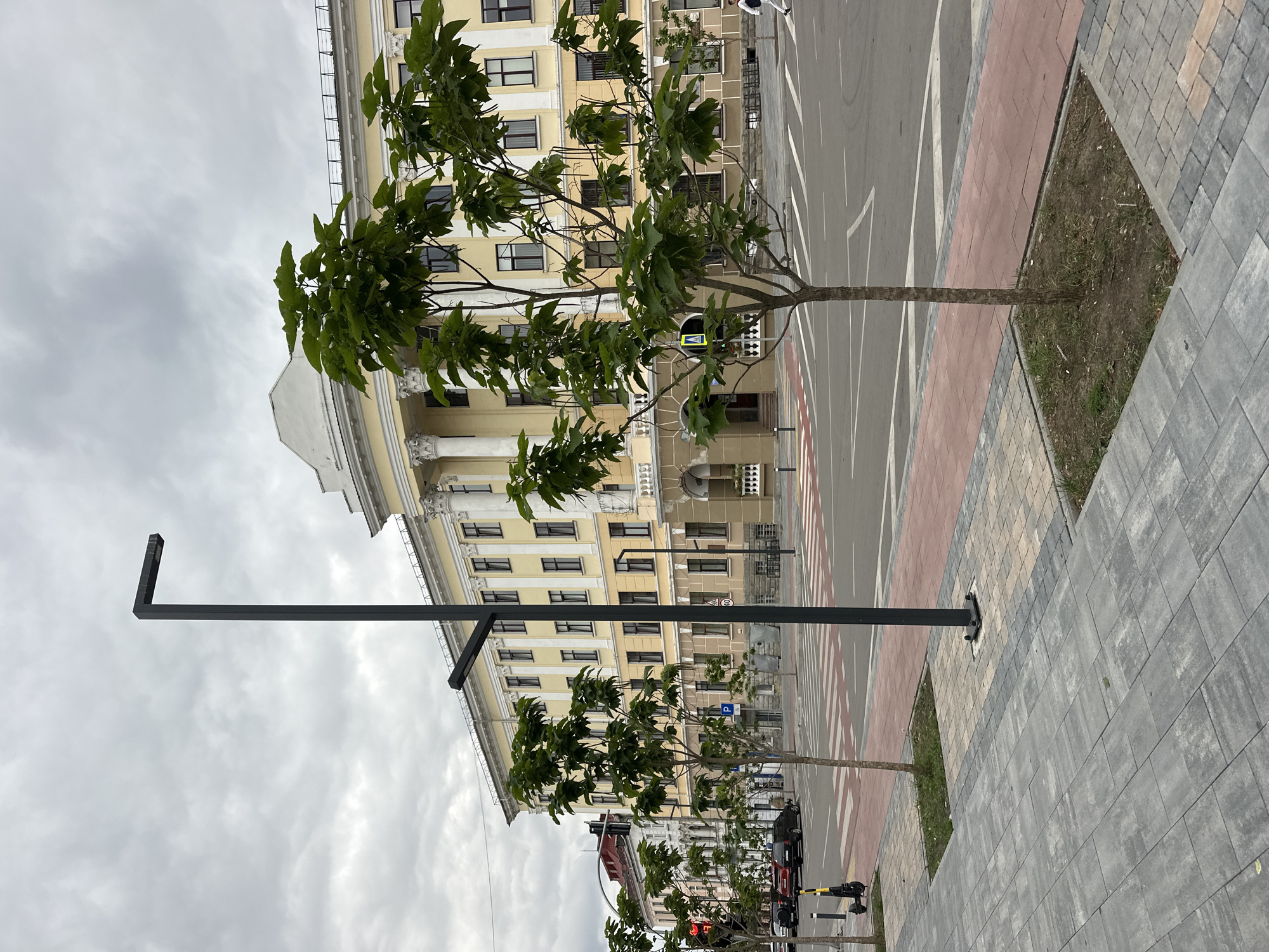 The image shows a street scene in Ternopil, Ukraine, featuring a large, yellow, multi-story building with classical architectural elements like columns and ornate trim. In the foreground, a modern dark grey lamppost stands on a paved sidewalk. Several young trees with green leaves are planted in square beds along the sidewalk, bordering a crosswalk marked with red and white stripes. Across the street, there are cars, including a black sedan and a red vehicle, and a yellow electric scooter is parked on the sidewalk. The sky is overcast with grey clouds.