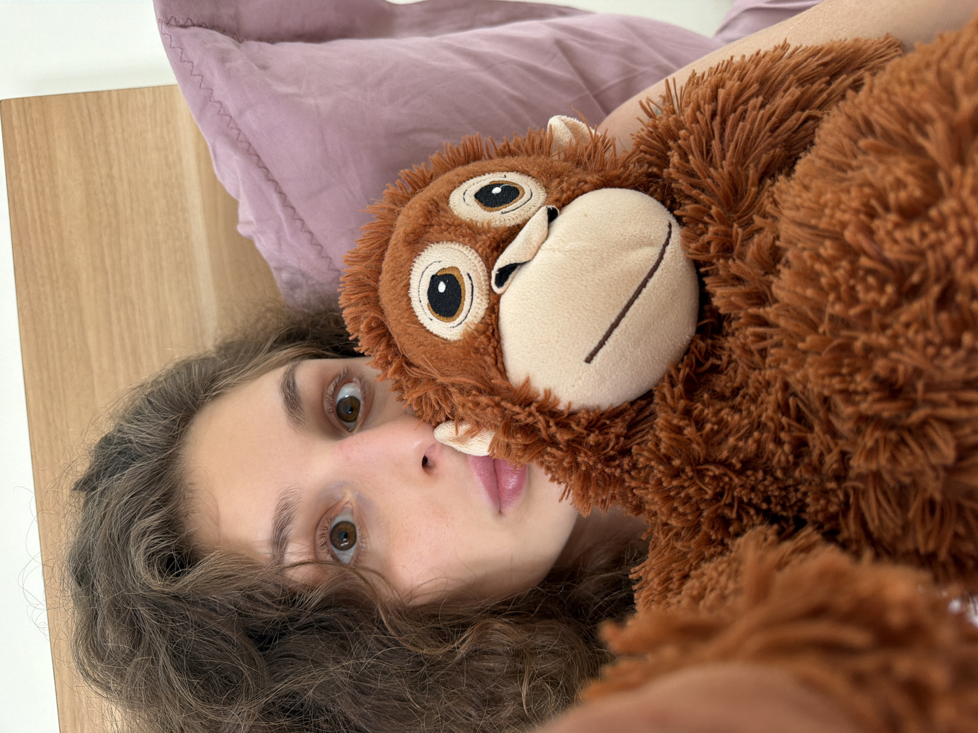 A fair-skinned young woman with brown, curly hair and light brown eyes lies on her back on a bed. She is holding a brown, plush monkey toy close to her face. Her head rests on a light purple pillow. A light-colored wooden headboard is visible to the left. The location is not discernible as North Kuta, Indonesia based on the visual information provided.
