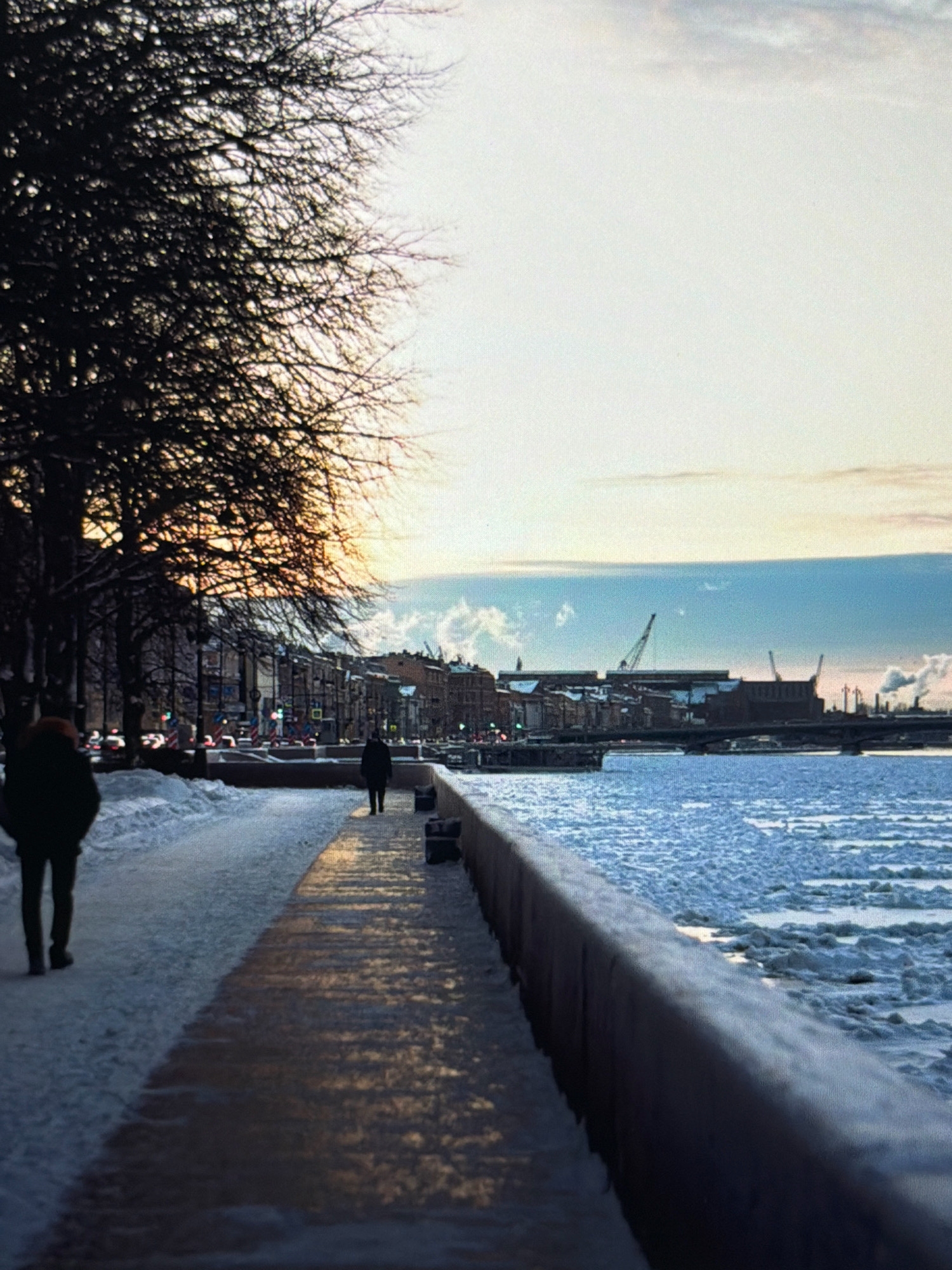 The image depicts a winter scene along a waterfront promenade. On the left, a leafless tree with dark branches dominates the foreground, casting a shadow. A person is walking away from the camera on a snow-covered path to the left of the promenade. The promenade itself is partially covered in snow and ice, with patches of wet pavement reflecting the low sunlight.

To the right of the promenade, the water is frozen or partially frozen, with large chunks of ice floating on the surface. A stone wall lines the edge of the promenade, separating it from the water. In the background, a city skyline is visible with buildings and industrial structures, including cranes and smokestacks emitting steam. The sky is overcast with a gradient of light colors, suggesting either sunrise or sunset. The overall atmosphere is cold and serene.

This image does not depict the city of Kafr Yasif, Israel. The architecture and industrial elements in the background suggest an urban environment in a colder climate, possibly a port city in Northern Europe.