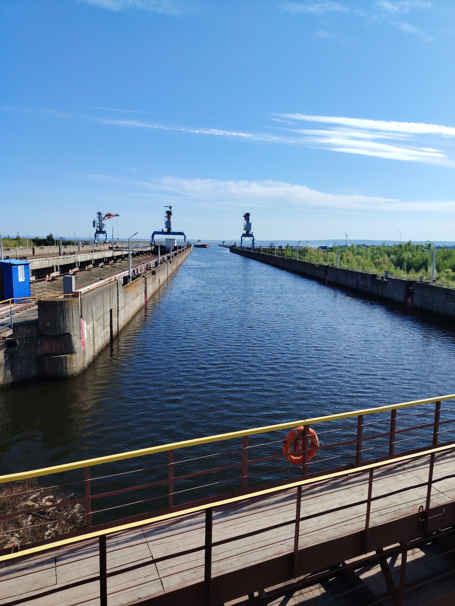 The image depicts a waterway, likely a canal or lock system, with concrete walls on either side. There is water in the canal. Three tall cranes are visible on the left. A small blue building sits next to the first crane. There is a dock with a wooden walkway extending from the bottom right of the image. An orange life preserver is attached to the railing. In the background, there is a line of trees on the far side of the water. The sky is bright blue with sparse white clouds. Given the location context, the image shows the Kama river and possibly the locks system near the city of Chaikovsky, Russia.