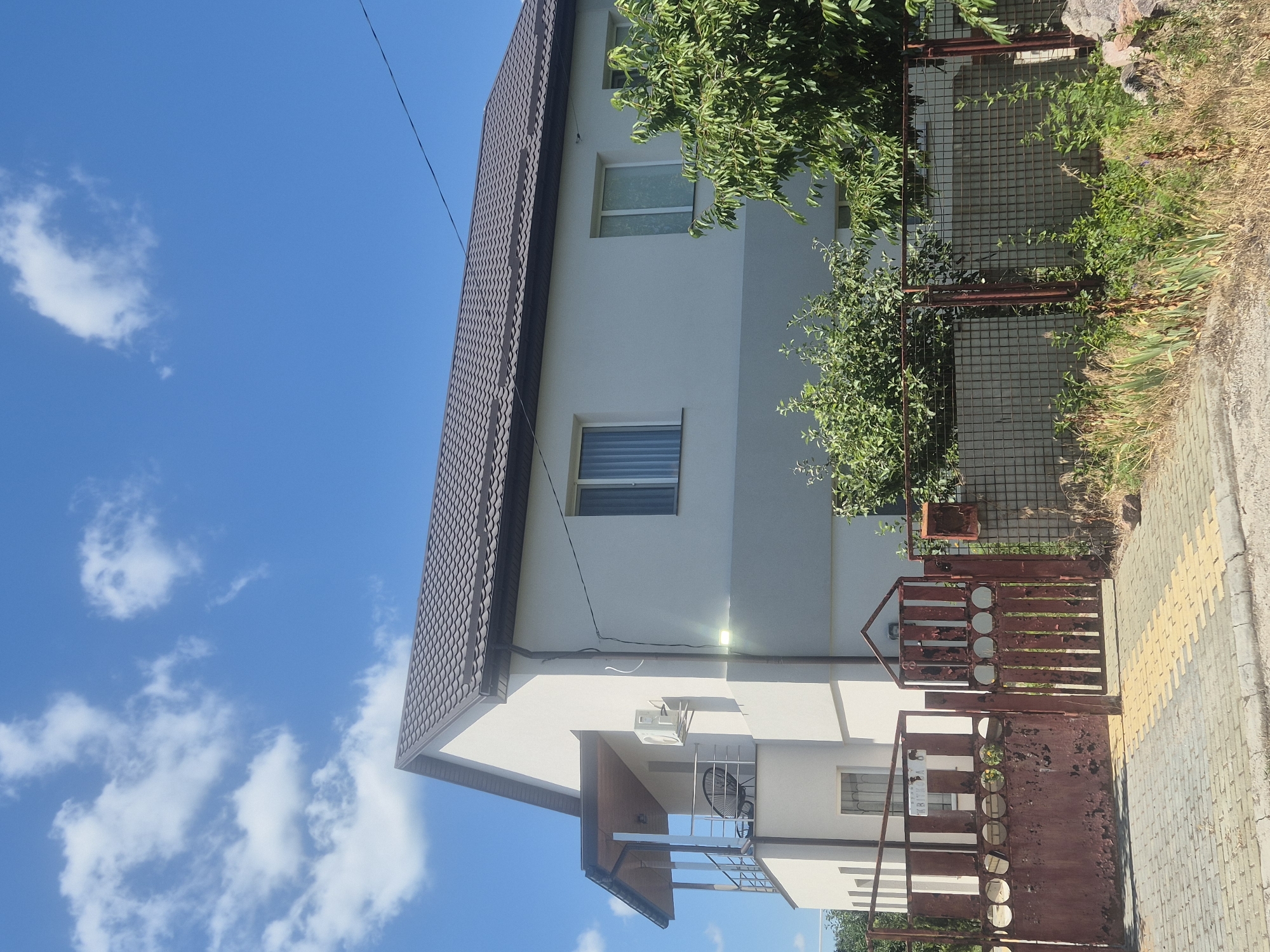 This is a full, eye-level, outdoor, vertical shot of a two-story house and its fence on a sunny day in the city of Sozonivka, Ukraine. The house is white with a brown metal roof and has a balcony with a railing. There are several windows on the second floor, and an air conditioning unit is mounted on the side of the house. In front of the house is a weathered, red metal gate and a chain-link fence. Lush green trees and overgrown vegetation are visible throughout the scene, and the sky is a clear blue with scattered white clouds.