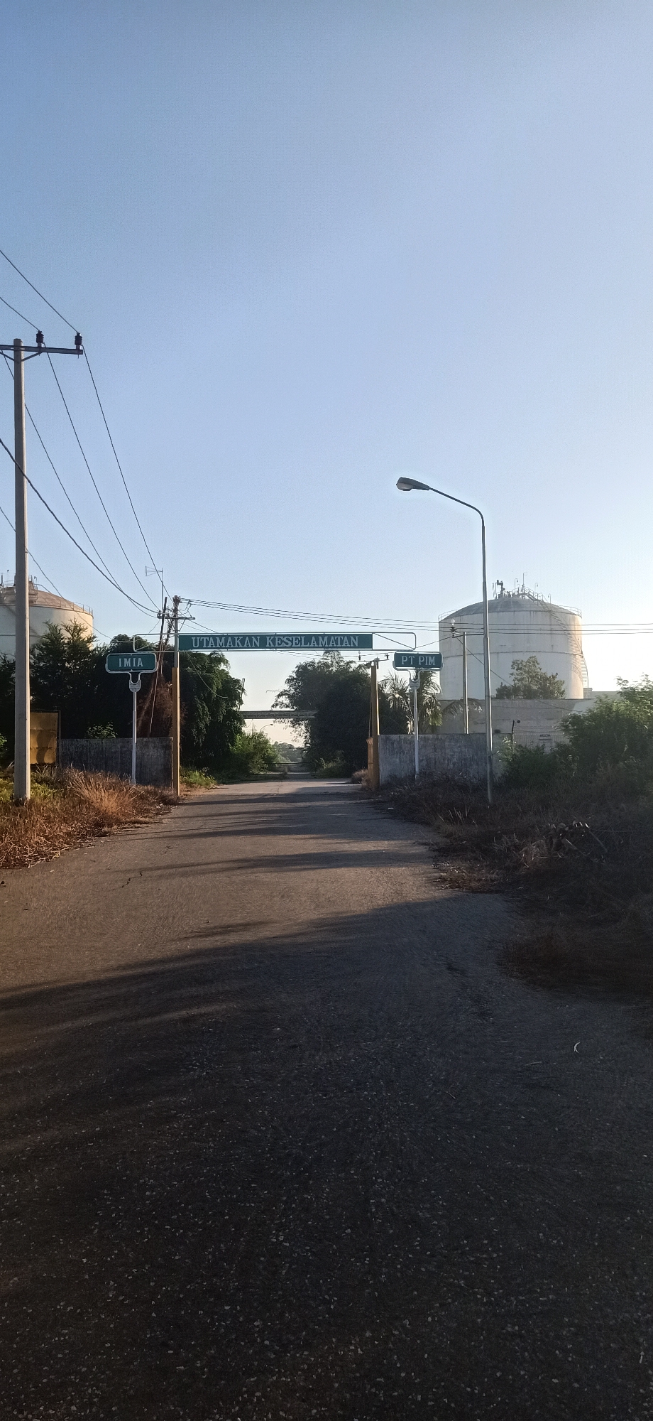 A clear, bright, daytime view depicts an asphalt road leading into what appears to be an industrial area in Keudekruenggeukueh, Indonesia. The sky is a pale blue, suggesting morning or late afternoon. On the left side of the road, a utility pole with wires stands tall, and a large, white cylindrical storage tank is visible behind it. On the right side of the road, another similar storage tank is partially obscured by foliage.
Two green signs are mounted on poles across the road. The sign on the left reads "IMIA" in white text. The central, larger sign reads "UTAMAKAN KESELAMATAN" in white text, which translates to "Prioritize Safety." The sign on the right reads "PT PIM" in white text.
The road itself is dark asphalt, showing some signs of wear and tear. Shadows from the utility pole and signs stretch across the road. The environment is a mix of dry, grassy vegetation and lush green trees. The scene is quiet and appears uninhabited.