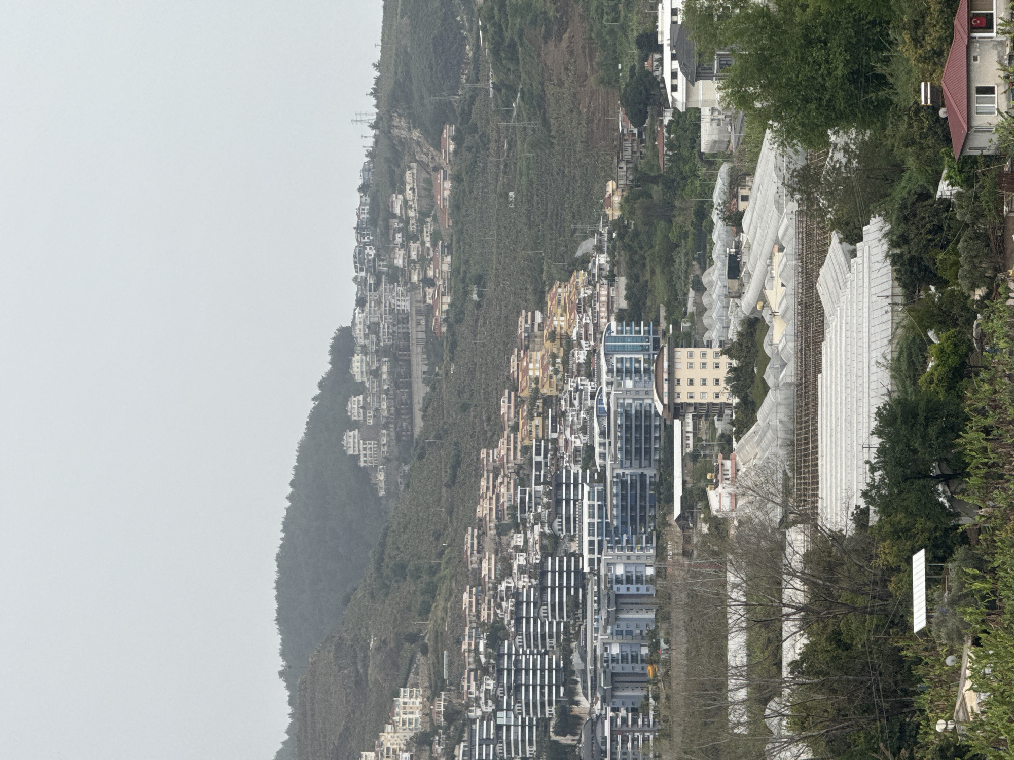 The image displays a densely populated hillside under an overcast sky, viewed from a slightly elevated perspective. In the foreground, several large structures with white, arched or sloped roofs, resembling greenhouses or industrial buildings, are prominent. Beyond these, a cluster of modern, multi-story buildings, characterized by their rectangular forms and numerous windows, line a street or main thoroughfare. One blue-toned building in this cluster has the word "marine" partially visible on its side.
Further up the steep hillside, a multitude of residential buildings, including houses and smaller apartment blocks, are tightly clustered. These structures exhibit varied colors, including white, beige, and yellow facades, and are interspersed among dense vegetation and trees. The upper reaches of the hill are heavily forested, with some power lines and utility poles visible traversing the landscape.
A notable detail is a small Turkish flag, red with a white star and crescent, prominently displayed on the side of a building with a red-tiled roof, situated in the mid-ground towards the right. The sky is a uniform, hazy light gray, indicating a heavily overcast day, which softens the light and creates a subdued atmosphere. No people are visible, and no specific activity or event is taking place, presenting a static panorama of a developed hillside environment.