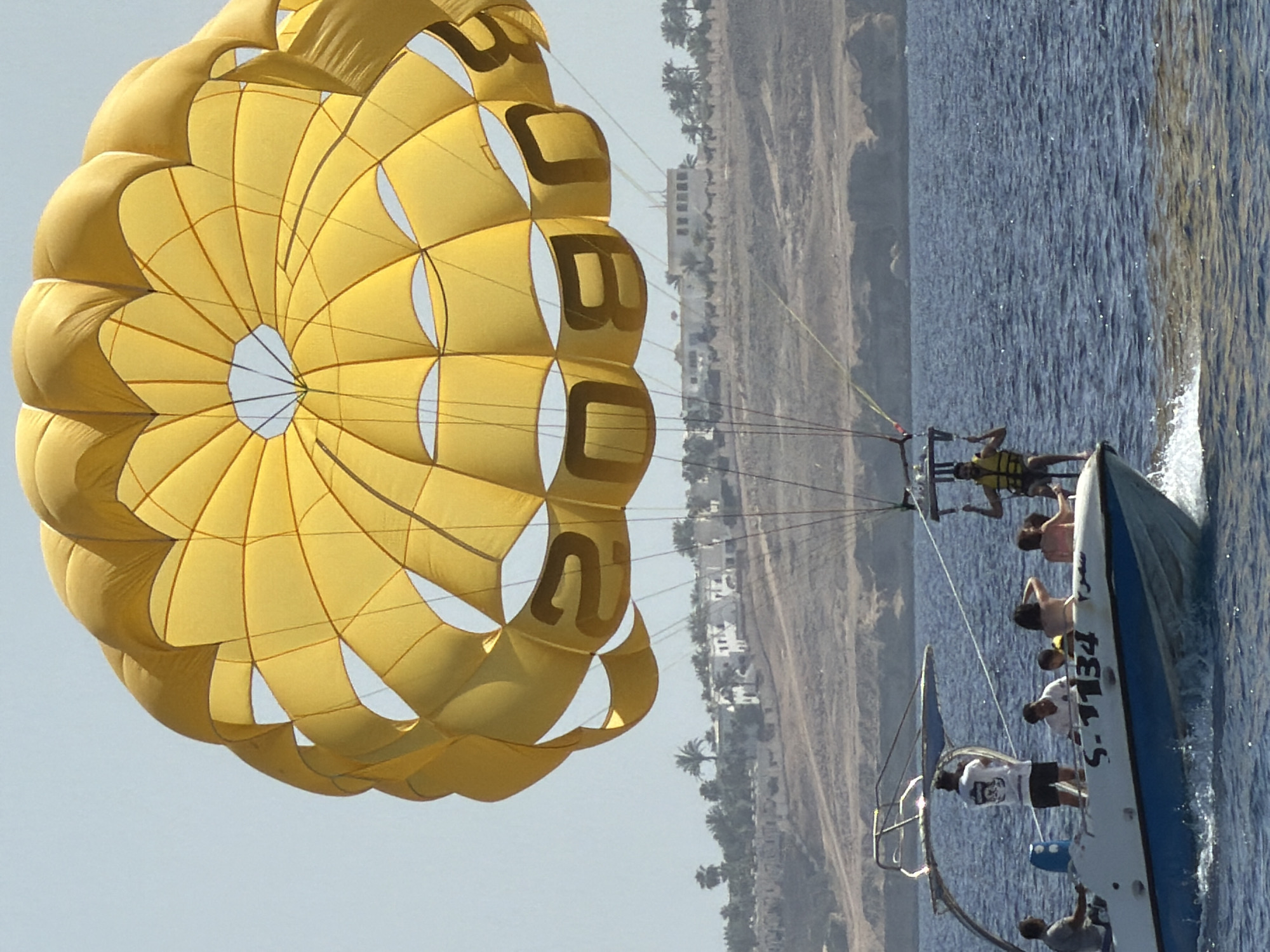 A parasailer is soaring over the Red Sea near Sharm El Sheikh, Egypt, being towed by a boat with several people on board. The large, yellow parachute is emblazoned with black lettering. The shoreline, dotted with palm trees and buildings, is visible in the background. The sea is choppy.