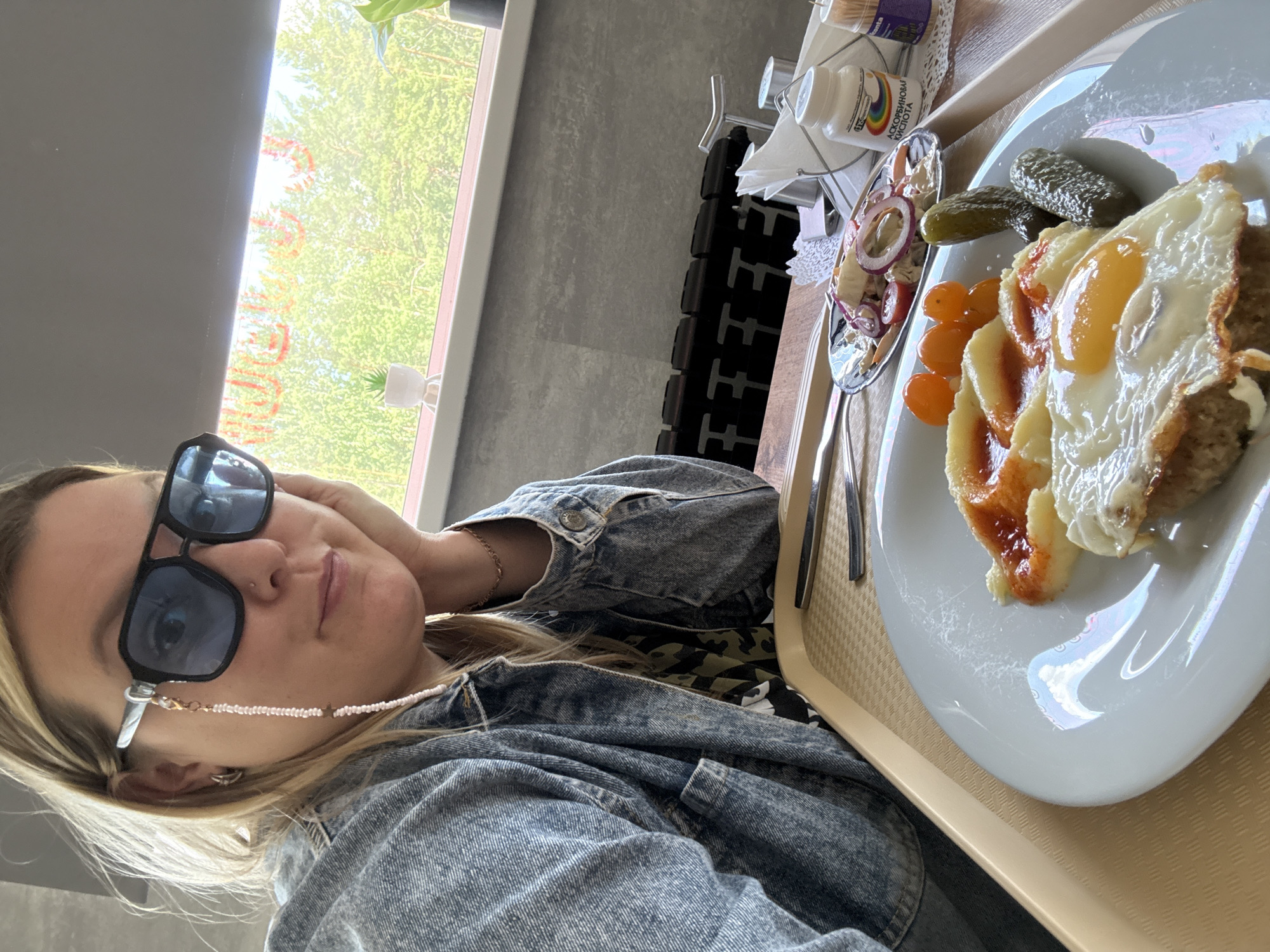 A woman with blonde hair is seated at a table, resting her head on her hand and looking towards the camera. She is wearing blue-tinted sunglasses, a denim jacket, and a patterned shirt. In front of her is a tray with a plate of food, including a fried egg, mashed potatoes with sauce, cherry tomatoes, and pickles. A small side dish of salad with onions and tomatoes is also visible. Behind the food tray, a bottle labeled "АСКОРБИНОВАЯ КИСЛОТА" (Ascorbic Acid) and toothpicks in a holder are present. The background shows a window with trees and foliage outside, and a radiator below the window. The environment appears to be an indoor dining area. The location is identified as Noviy, Russia.