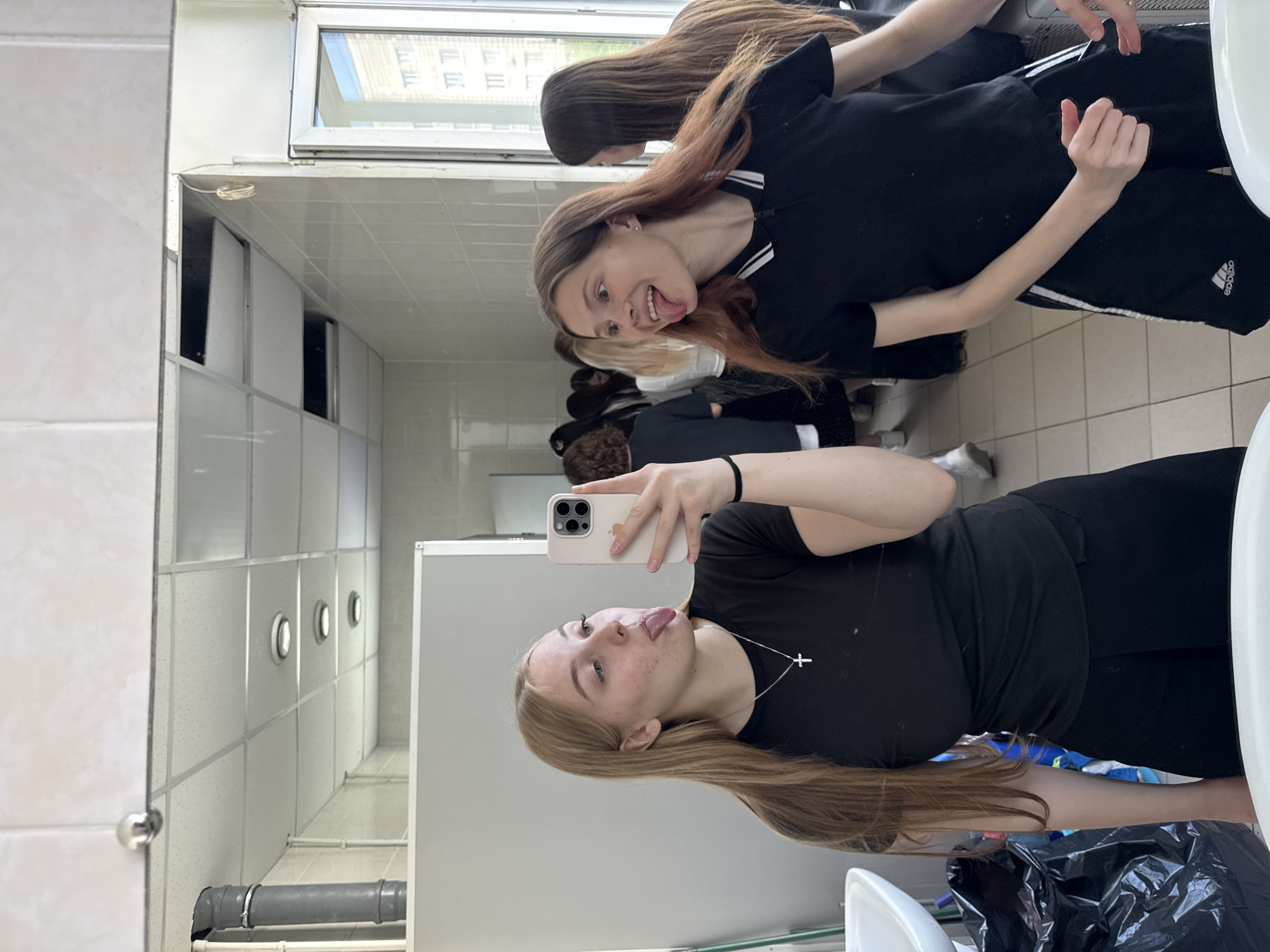 Two young women pose for a selfie in a bathroom. The woman in the foreground holds a cell phone and sticks her tongue out. She wears a black shirt and long silver necklace with a cross pendant. The second woman in the background also sticks her tongue out and wears a black shirt with white trim and black athletic pants with white stripes. The bathroom has a tiled ceiling and walls with small recessed circular lights. In the background, there are other people in the bathroom. The location is Perm, Russia.