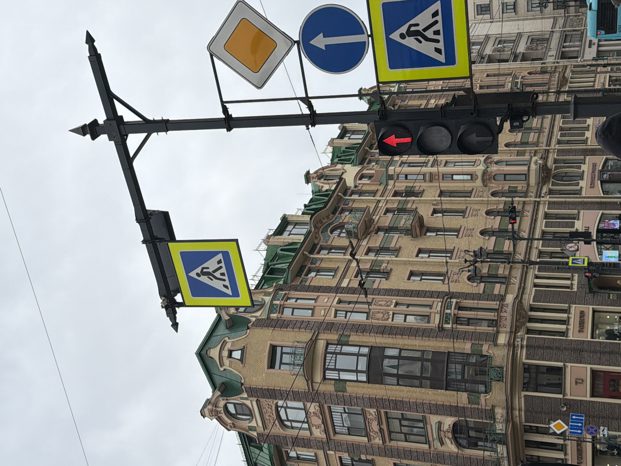 An outdoor, slightly askew, eye-level shot captures a street scene in Saint Petersburg, Russia, under an overcast sky. A prominent black metal pole, featuring decorative finials, spans horizontally across the upper half of the frame, supporting various road signs and a traffic light array.
From left to right on the pole, a square blue pedestrian crossing sign with a white figure walking is visible. Further right are a yellow diamond-shaped 'main road' sign and a blue circular sign with a white left-pointing arrow, indicating a mandatory left turn. Below these, a traffic light displays a red left-turn arrow, suggesting vehicles in that lane must stop or cannot turn left. Another pedestrian crossing sign is located further to the left, lower on the pole structure.
The background is dominated by a multi-story, ornately designed building, characteristic of historic European architecture with elements consistent with Saint Petersburg. The building features light brown brickwork, numerous windows, some with intricate balconies, and decorative green roofs or cornices. Smaller street signs, including another yellow diamond 'main road' sign, a blue 'no stopping' sign, and a blue 'mandatory right turn' sign, are affixed to the lower part of the building.
Visible text includes "TJ" and "НОВА" (NOVA) on storefronts or advertisements on the building. A bus with the number "6753" is partially visible in the upper right background. The image depicts a daytime setting with diffused lighting due to the cloudy weather.