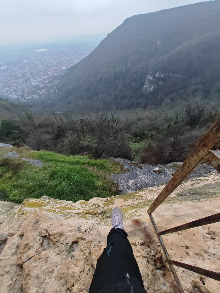 The scene is a first-person, downward-angled view from a rocky precipice overlooking the city of Shumen, Bulgaria. In the immediate foreground, a person's right leg, clad in black outdoor pants with a zippered pocket and a light grey athletic shoe, extends over a textured, light-colored limestone ledge, which exhibits patches of yellow-green lichen. To the right, a section of a rusty, dark-colored metal ladder or railing, constructed from square-sectioned bars, descends partially out of view. Below the ledge, the steep slope transitions into areas of vivid green vegetation and dark, exposed rock, leading to a dense, predominantly bare deciduous forest that covers the valley floor and the opposing mountain flank. The city of Shumen is visible in the distant background, characterized by a dense arrangement of structures with reddish roofs, situated in a valley beneath an overcast grey sky. A lighter, flat expanse stretches beyond the city to the horizon.