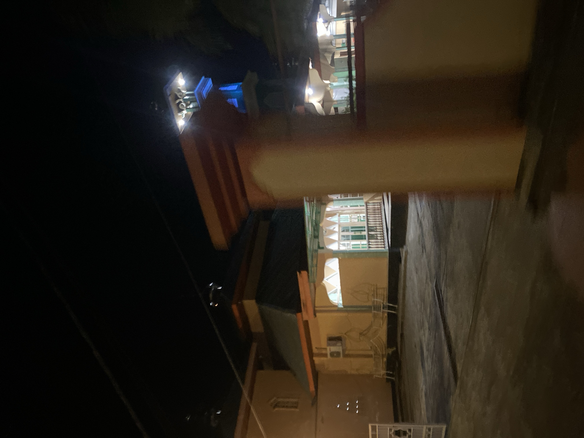 This is a night-time photograph of a building with a distinctive tower, likely a mosque or religious building, in Bantaeng, Indonesia. The building is illuminated by artificial lights, casting shadows and creating a somewhat hazy atmosphere. The foreground is dominated by a blurry, out-of-focus orange and yellow structure, possibly a wall or pillar, which suggests the photograph was taken from a moving vehicle or by someone in motion.
To the left, a tan-colored building with a dark sloped roof is visible. It features a prominent air conditioning unit on its wall and a series of small, circular lights above a window. White benches are arranged in front of this section of the building. Further to the right, an open-sided structure with a pitched roof and white window frames is partially obscured.
The prominent tower, which is illuminated with blue light at its upper section, rises above the main building. The sky is dark, indicating it is nighttime, and several power lines are visible crisscrossing the upper portion of the frame. The ground appears to be paved. The overall impression is that of a public building observed at night.