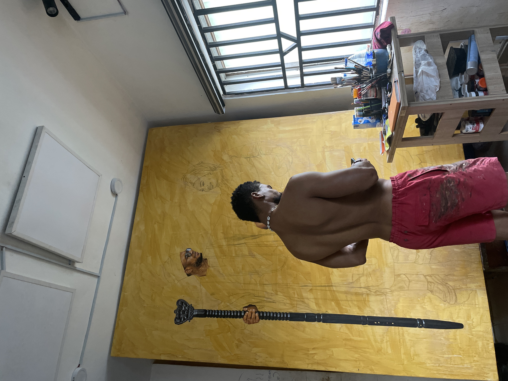 An artist is captured in a brightly lit studio, working on a large, vertical canvas that dominates the scene with its textured golden-yellow background. The image is taken from a slightly low angle, looking up. The artist, a shirtless man with dark skin, short dark hair, and a shell necklace, is seen from behind, facing the canvas. He wears red shorts, which are smudged with paint, indicating he is actively engaged in his craft.
On the canvas, preliminary sketches in light pencil are visible, including the outline of a figure on the upper right and possibly another face on the left. More prominently, a painted head of a man with glasses and a beard is discernable on the lower left, along with a painted hand firmly grasping a black, ornate ceremonial staff or scepter. The staff extends downwards toward the bottom of the canvas.
To the left, a stack of white square panels or canvases leans against the wall, alongside circular light fixtures. Above, a large skylight with black metal grates provides ample natural light, illuminating the workspace. To the right of the artist, a wooden shelf unit is overflowing with various art supplies, including numerous paintbrushes, tubes of paint, bottles, and other tools, suggesting a vibrant and active creative environment. The walls are light-colored. The scene depicts a focused moment in an artist's studio, likely in Ado, Nigeria, during daylight hours. No discernible text is visible in the image.