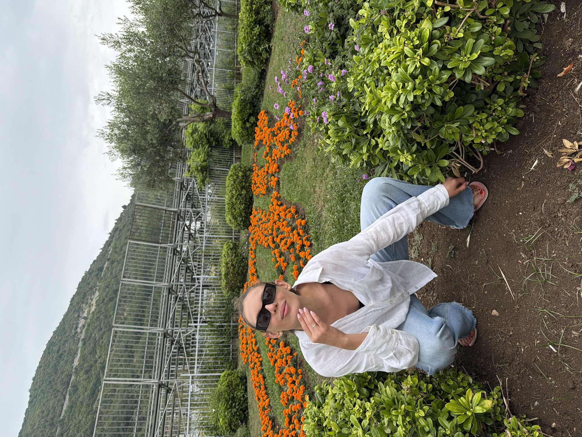 A woman is captured in a squatting or kneeling pose on a dirt path, looking towards the camera. She is wearing dark sunglasses, a loose white long-sleeved shirt, and light blue jeans. Her hair is pulled back, and she has a ring on her right hand, which is resting near her chin.
The setting is an outdoor garden area. To the woman's immediate left and in front of her are vibrant orange flowers, likely marigolds, arranged in beds, and various green bushes. Some smaller purple flowers are also visible among the greenery. The ground is a mix of green grass and dark soil.
In the mid-ground, a large, intricate metal structure, resembling scaffolding or a framework for a greenhouse, extends across the scene. Beyond this structure, a lush, tree-covered mountain dominates the background, indicating a hilly or mountainous landscape. The sky is overcast, suggesting a cloudy day with soft, diffused light. An olive tree with sparse foliage stands on the left side of the frame. The overall atmosphere appears calm and natural. There is no visible text in the image. The location is contextualized as Budva, Montenegro.