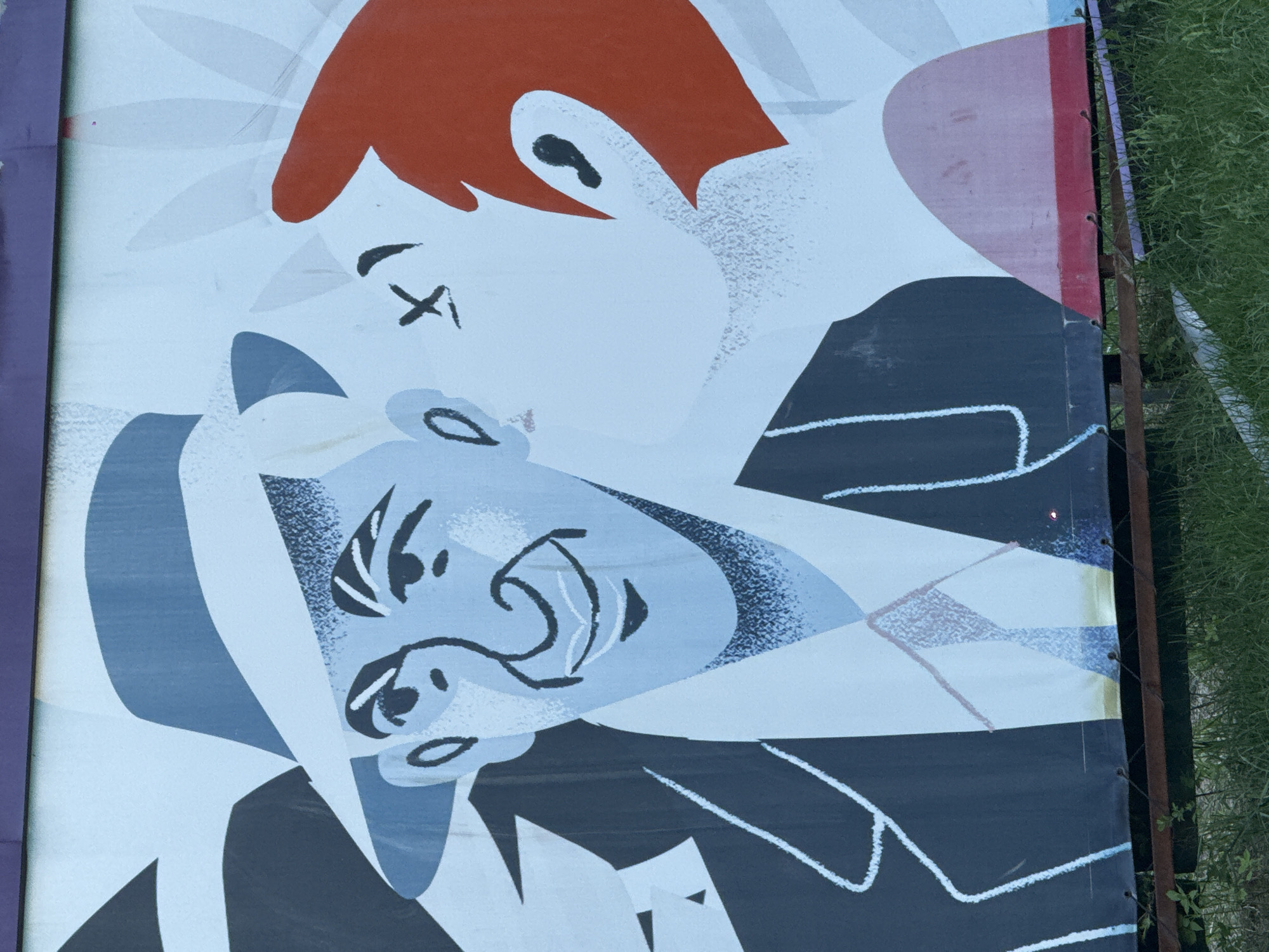 An outdoor poster in Moscow, Russia, depicts two men embracing. The man on top has short, orange hair and a white face with a black "X" for an eye. He wears a dark suit outlined in white. The man below has bluish skin and dark hair, also wearing a dark suit. The poster is attached to a metal frame, with grass visible to the right. The left side of the poster has a purple stripe.