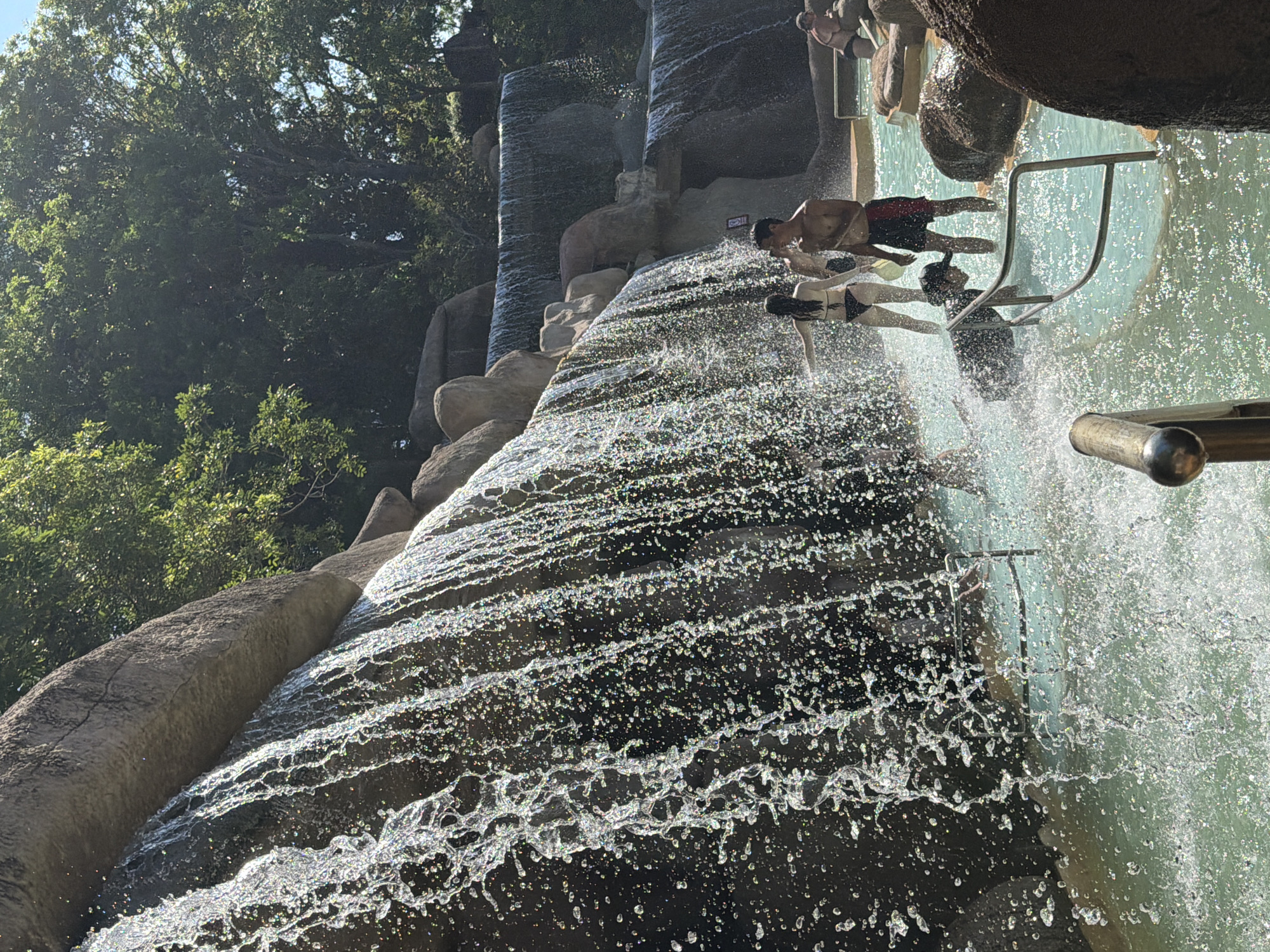 The image depicts a scene at a water park, likely located in Tay Nha Trang Ward, Vietnam. The primary feature is a multi-tiered artificial waterfall cascading down dark rock formations. Several people are visible interacting with the water. In the foreground, a person wearing a black bikini stands facing away from the camera, with water spraying around them. To their right, a man in red and black swim trunks stands, looking towards the waterfall. Further to the right, another man in swim trunks is seated on a rock. In the middle ground, a younger person wearing a dark top and hat is partially submerged in the water, holding onto a metal railing. The water itself is a pale turquoise color and appears to be a pool or shallow waterway. The background consists of lush green trees and more of the rock-like structures of the waterfall. The lighting suggests a bright, sunny day.