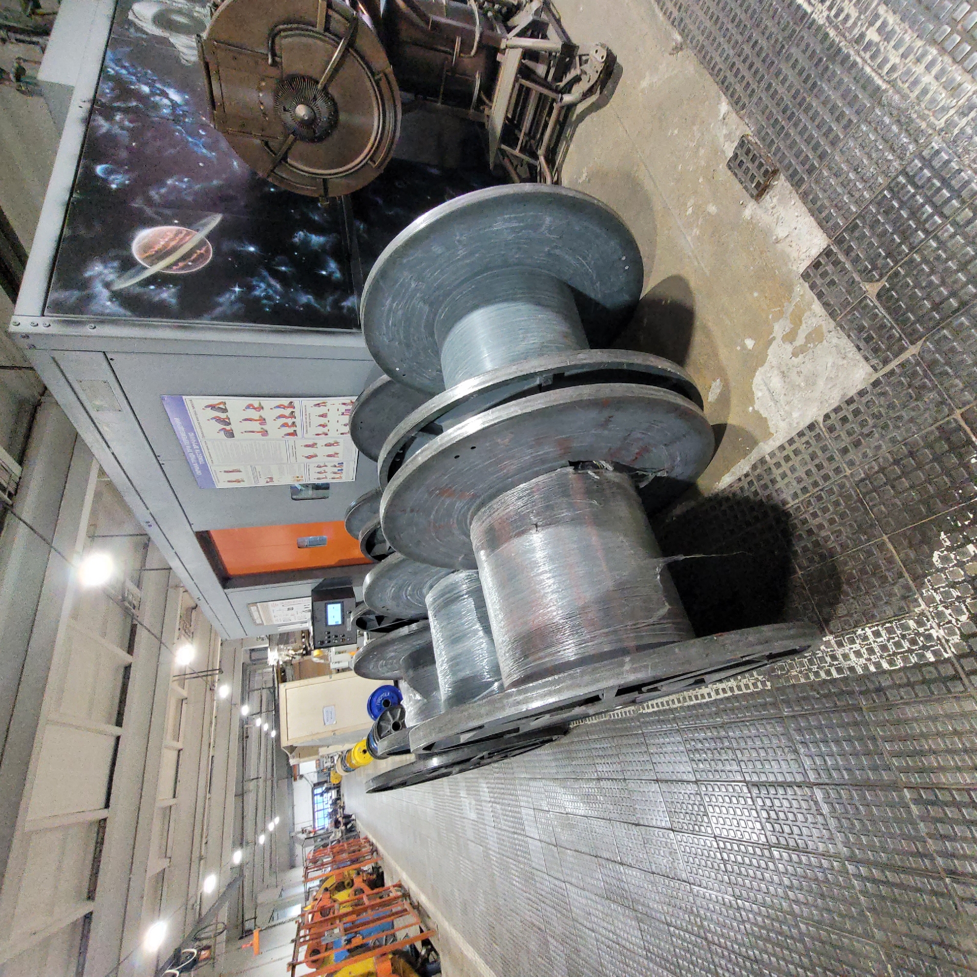 This image, taken in an industrial setting and rotated 90 degrees clockwise, captures a long factory floor or production line. In the foreground, several large, grey industrial spools or bobbins are lined up. These spools are wound with what appears to be wire or cable, most of which is neatly wrapped in clear plastic.
To the right, a textured metal surface, likely a floor or walkway, extends into the distance, characterized by a repeating pattern of small squares. To the left, a large piece of grey industrial machinery dominates the upper half of the frame. On one side of this machine, a vibrant graphic of outer space is displayed, featuring a planet resembling Saturn with its rings, alongside nebulae and stars. Above this graphic, another part of the machine includes a complex, circular component with spokes.
Below the space graphic, on a grey panel, a printed poster or chart is visible. It depicts various human figures in different poses, suggesting it might be an ergonomic guide or a safety poster with stretching exercises. Further down the line, beyond the spools, other industrial machinery with orange and yellow components can be seen. The background reveals high ceilings with rows of bright artificial lights and exposed industrial structures, indicating a large manufacturing or processing facility. No people are visible, but the arrangement of equipment suggests an active workspace.