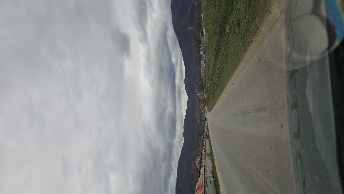 The image displays a scene in Kliment, Bulgaria, captured from within a vehicle, indicated by a reflection on the dashboard at the bottom. A grey asphalt road occupies the foreground and midground, extending towards a distant settlement. To the immediate left of the road, a low concrete wall precedes several residential-style buildings with light-colored facades and red-tiled roofs, some appearing unfinished. To the right, an expanse of green grass and dirt constitutes undeveloped land. Further along the road, a cluster of more distant buildings is visible, one of which prominently features a slender minaret with a conical cap. A large, dark-colored truck is observed moving away from the viewer on the road in the midground. The background consists of dark, undulating mountains or hills, covered with sparse vegetation. The sky is uniformly overcast with heavy, grey clouds, indicating dull or impending weather. No individuals are discernible.
