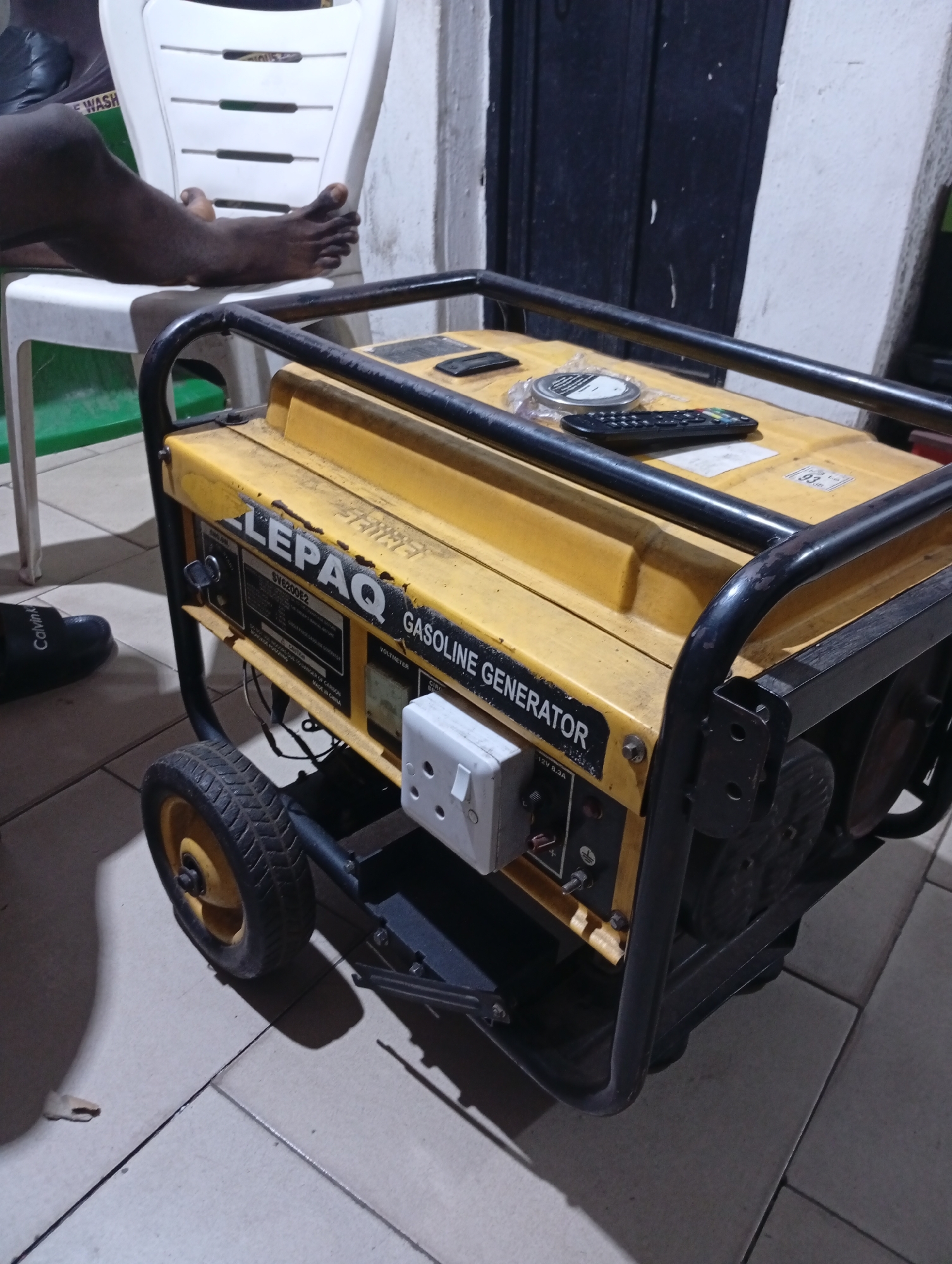 A yellow and black LEPAQ gasoline generator sits on a tiled floor. A white plastic chair with a dark-skinned person seated is visible in the background. A remote control and a cylindrical object are on top of the generator. A pair of black shoes are visible on the left. The location is Lokoja, Nigeria.
