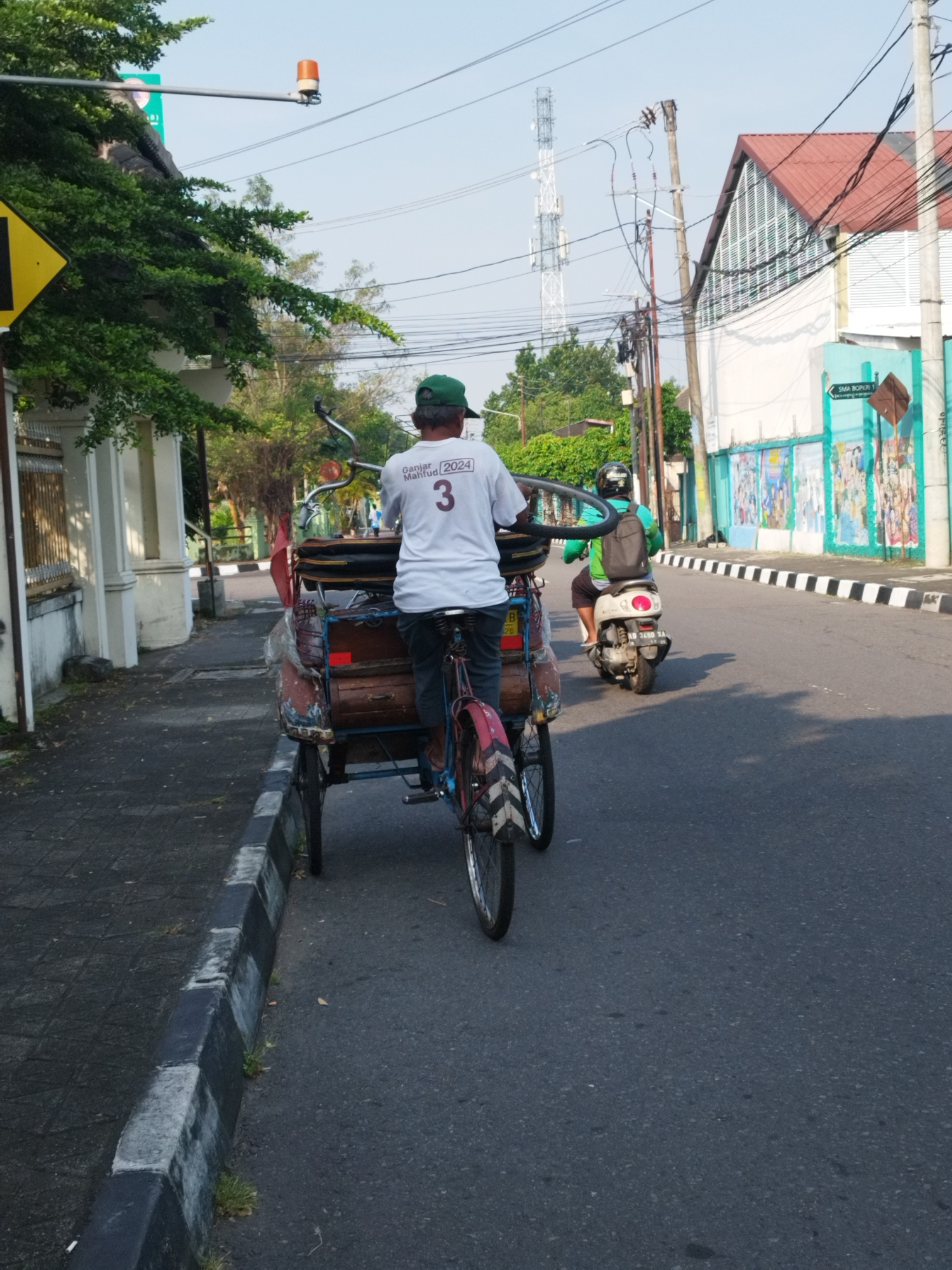 In the city of Yogyakarta, Indonesia, an overcast sky looms above a street scene. On the left, a yellow diamond-shaped road sign with a black border is partially visible, its reflection caught on a nearby building. A man, wearing a green cap and a white t-shirt with the text "Ganjar Mahfud 2024" and the number "3" printed on it, is seated on a becak, a three-wheeled passenger pedicab. The becak, laden with various items, is positioned on the side of the road, its front wheel turned towards the left. The man's hands are on the handlebars, suggesting he is either preparing to move or has just stopped. Further down the street, another individual, wearing a green shirt and a helmet, rides a white scooter, heading in the same direction as the becak. The scooter has a license plate that reads "KB 3499 XA". The street is lined with a black and white striped curb, separating the road from a sidewalk. Buildings with white walls and red tiled roofs are visible in the background, with a large tower, possibly a communication tower, standing tall against the sky. The overall impression is a glimpse into the everyday life and transportation methods of Yogyakarta.