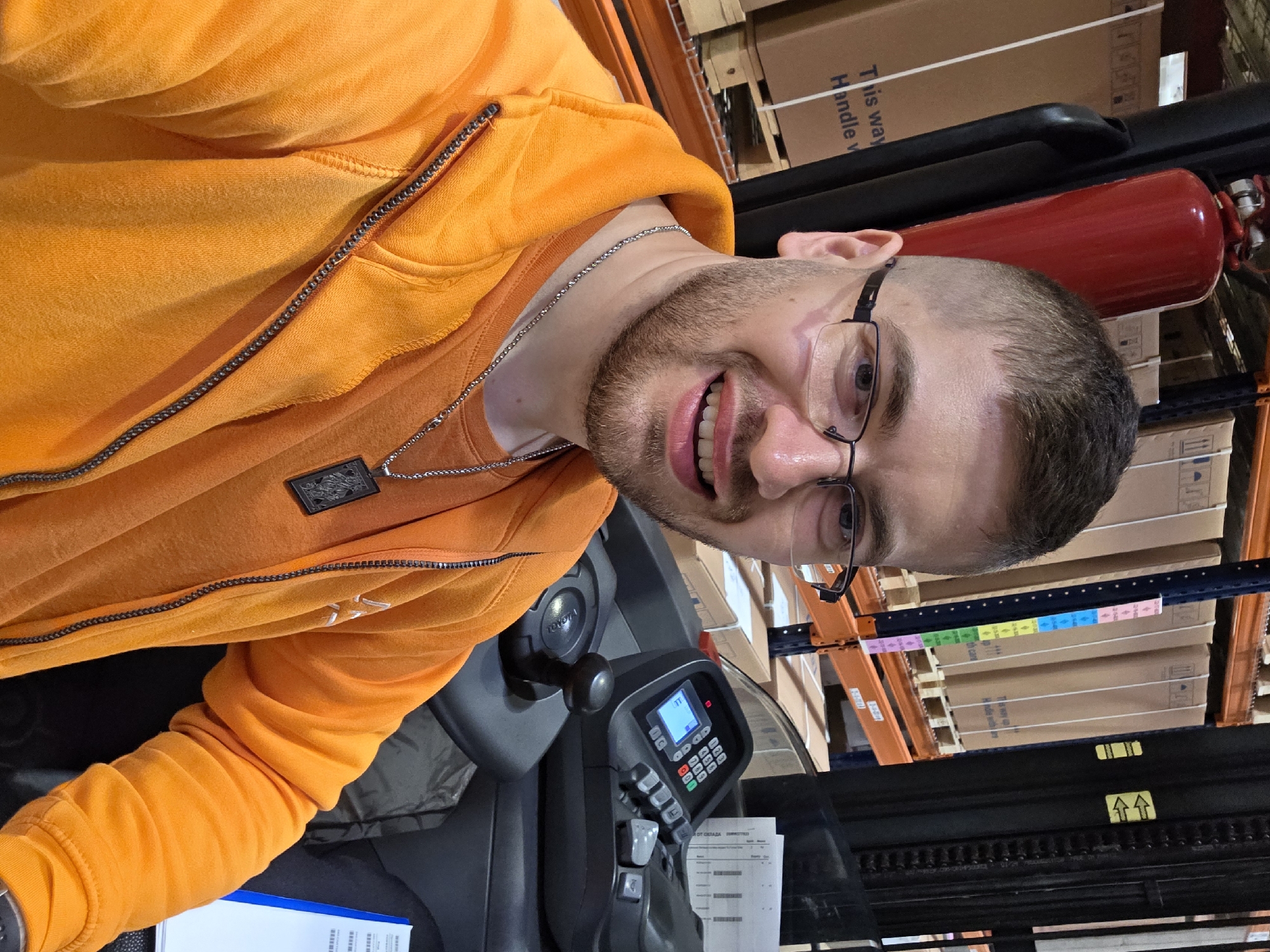 A man with a beard and glasses is seated in what appears to be the driver's seat of a forklift, possibly in a warehouse in Plovdiv, Bulgaria. He's wearing an orange hoodie and a silver chain. The forklift is gray, with a control panel visible. Behind him, shelves are stacked with boxes and a red fire extinguisher is visible. The background suggests a storage or distribution facility.