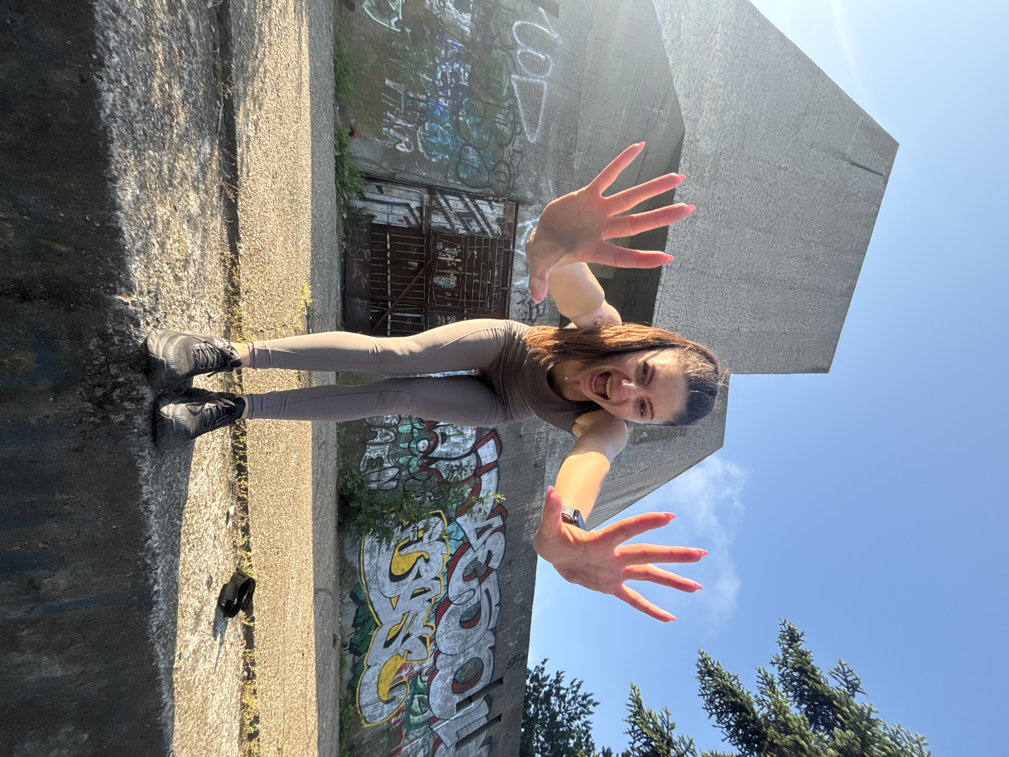 This image, originally rotated 90 degrees counter-clockwise, captures a dynamic outdoor scene on a bright, sunny day in Varna, Bulgaria. A young woman is the central subject, posing energetically. She has long brown hair pulled back, a wide, open-mouthed smile, and is wearing a fitted taupe-colored athletic top and matching leggings, paired with black athletic shoes. Both arms are extended forward towards the viewer with open palms, her fingers spread, and nails painted pink, suggesting a playful reach or push. A black smartwatch is visible on her left wrist.
She stands on a concrete surface, with strong sunlight casting clear shadows. Near her feet, a single black flip-flop lies on the ground. Behind her is a large, weathered concrete structure, possibly a building or wall, extensively covered in colorful graffiti. Visible text within the graffiti includes stylized white letters with yellow outlines forming what appears to be "GOOD," along with another white tag resembling "LOL" higher up, and various other illegible tags in white, blue, and green. A dark, rusty metal gate or door is embedded within this graffiti-covered concrete. To the right of the frame, the tops of green evergreen trees are visible against a clear, bright blue sky with a few wispy clouds. The strong shadows and vibrant light indicate it is daytime, likely midday or early afternoon. The overall mood conveyed is one of joy and energy.