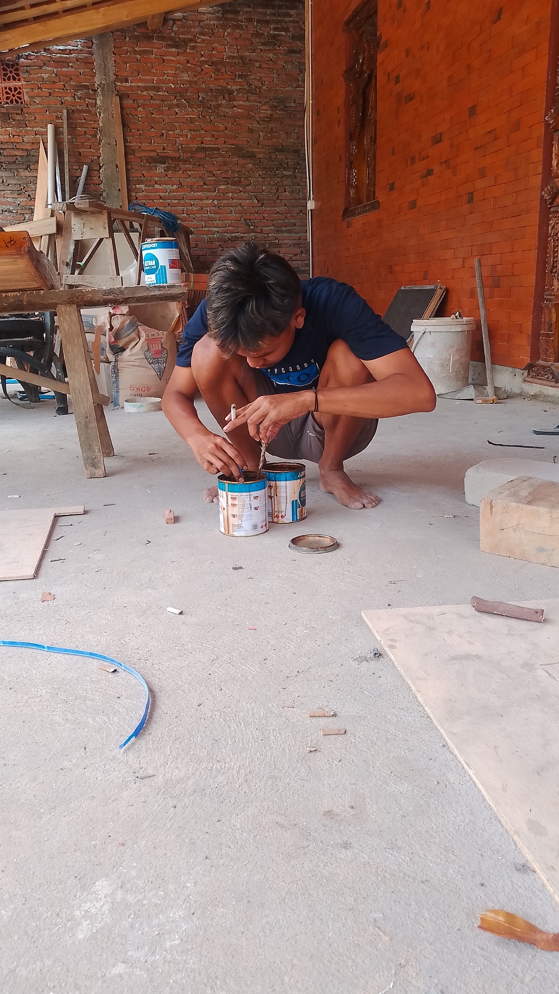 An Indonesian man is squatting on a concrete floor, working with two cans of what appears to be wood stain or paint. He is holding a stick in his right hand, stirring the contents of the can on his left. A third can, open, rests on the ground to his right. The man is wearing a dark blue t-shirt and shorts. The environment suggests an outdoor workspace, possibly a garage or workshop, with brick walls, wooden structures, and construction materials like plywood and bags of what looks like cement. The location is identified as Karanglangu, Indonesia.