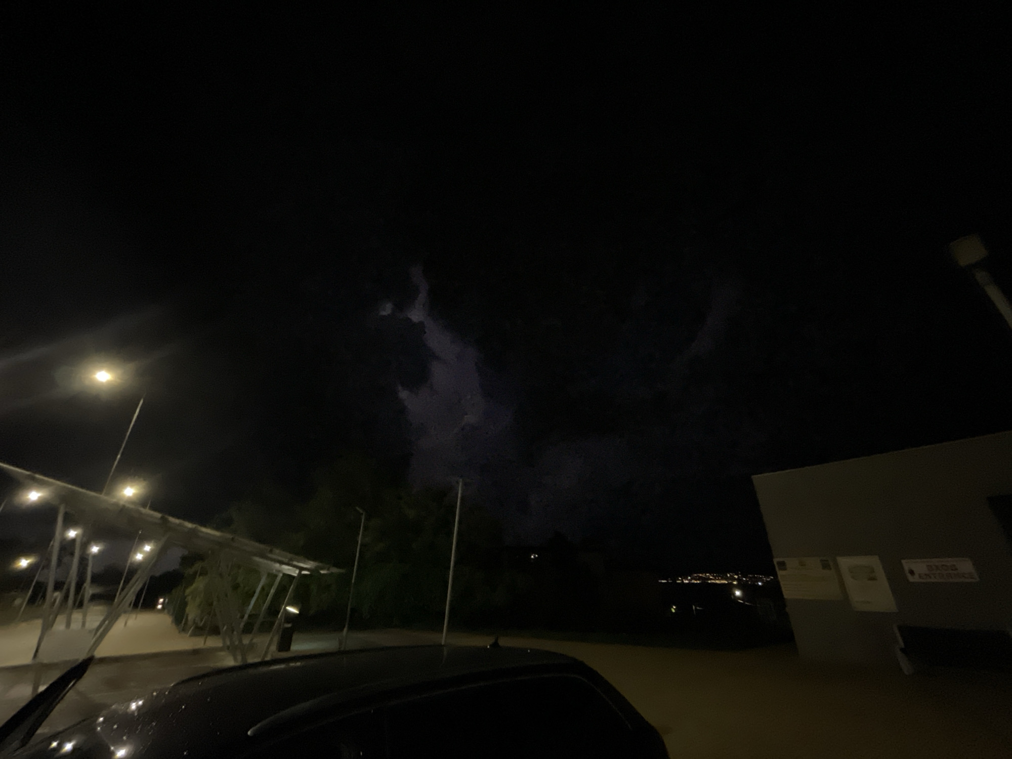 A nighttime scene showing a car parked near a building with a sign reading "EKOS ENTRANCE." In the background, the lights of a city can be seen, likely Byala, Bulgaria. The sky is dark with what appears to be either storm clouds or a lightning strike visible in the upper center of the frame. Streetlights illuminate a covered walkway to the left.