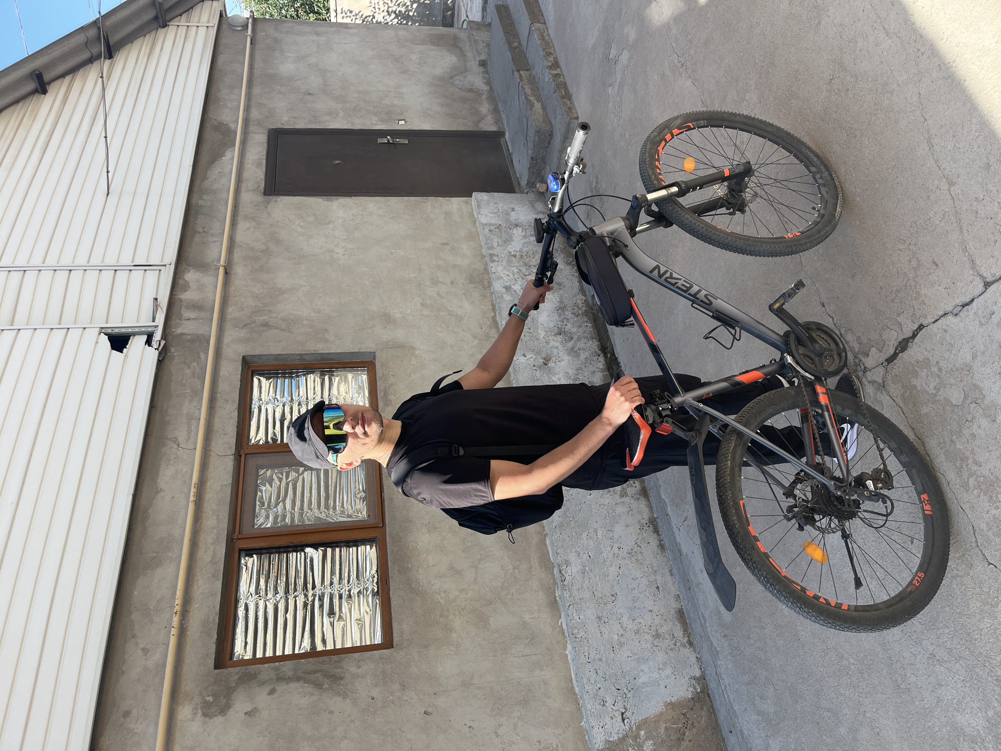 A man wearing a black t-shirt, black pants, a black baseball cap, and bright, multi-colored sports sunglasses stands next to a dark gray and orange mountain bike. He has a black backpack on his shoulders. The man is holding the handlebars of the bicycle. The bicycle has a black saddle with orange accents and a black frame bag. The scene appears to be in a residential area with a concrete building in the background. The building has a grey concrete facade, a white vinyl-sided upper portion with some damage near a window, and a dark brown door. There are two windows on the concrete portion of the building, covered with reflective material. A concrete staircase leads up to the door. The ground is paved with concrete. The image was taken on a sunny day. The man's smartwatch is visible on his left wrist. The bicycle has the brand name "STERN" on its frame. The rear tire is labeled "27.5". The location is the city of Shymkent, Kazakhstan.