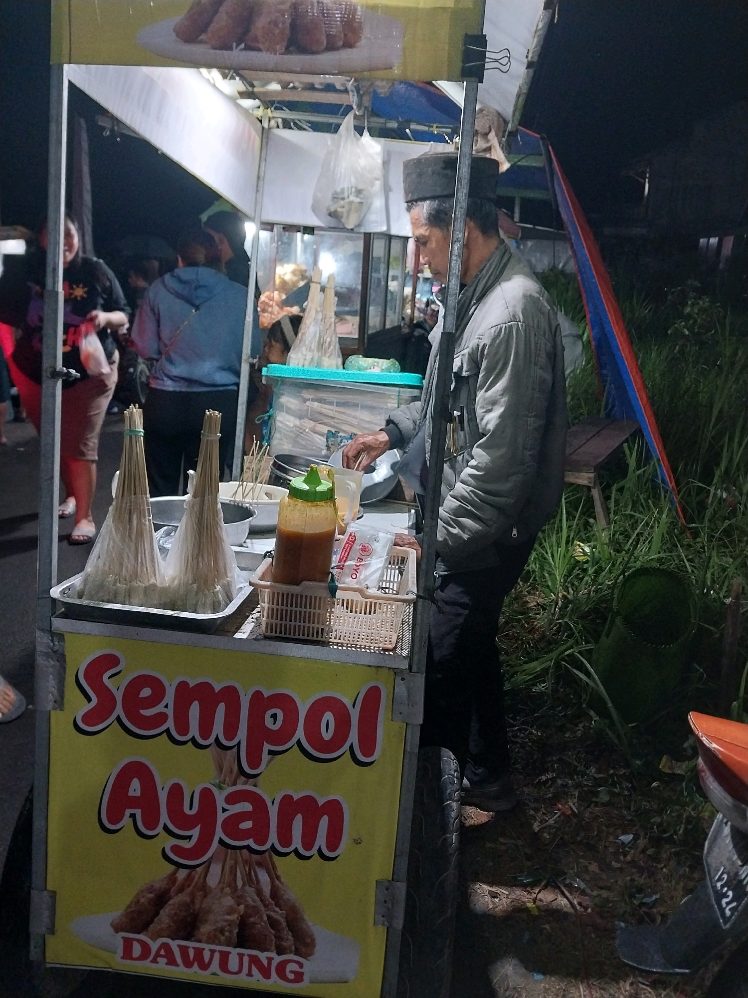 A nighttime street food vendor scene is depicted in Nglegok, Indonesia. A man, appearing to be the vendor, stands behind a food cart. He is wearing a dark cap and a gray jacket. The cart has a yellow banner with red lettering that reads "Sempol Ayam DAWUNG" and features an image of fried food on skewers. In front of the cart, a tray holds two bundles of skewers wrapped in plastic. To the right of the skewers is a basket containing packaged food items and a bottle of orange sauce. Behind the cart, other people are visible, including a woman wearing a black t-shirt and holding a plastic bag. The environment appears to be a roadside, with tall grass and a hint of a motorcycle on the right side of the frame. The lighting is dim, suggesting it is nighttime.