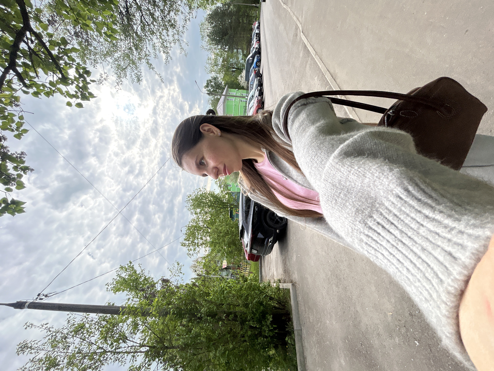 A daytime, ground-level, medium shot presents a woman in Klin, Russia. She is outside, standing on a paved surface, possibly a street or sidewalk, with buildings and parked cars in the background. The woman is wearing a light grey sweater over a pink shirt. A dark brown handbag hangs from her shoulder. Trees with green foliage are visible around the area, and a grey, cloudy sky is above. A utility pole is on the left edge.