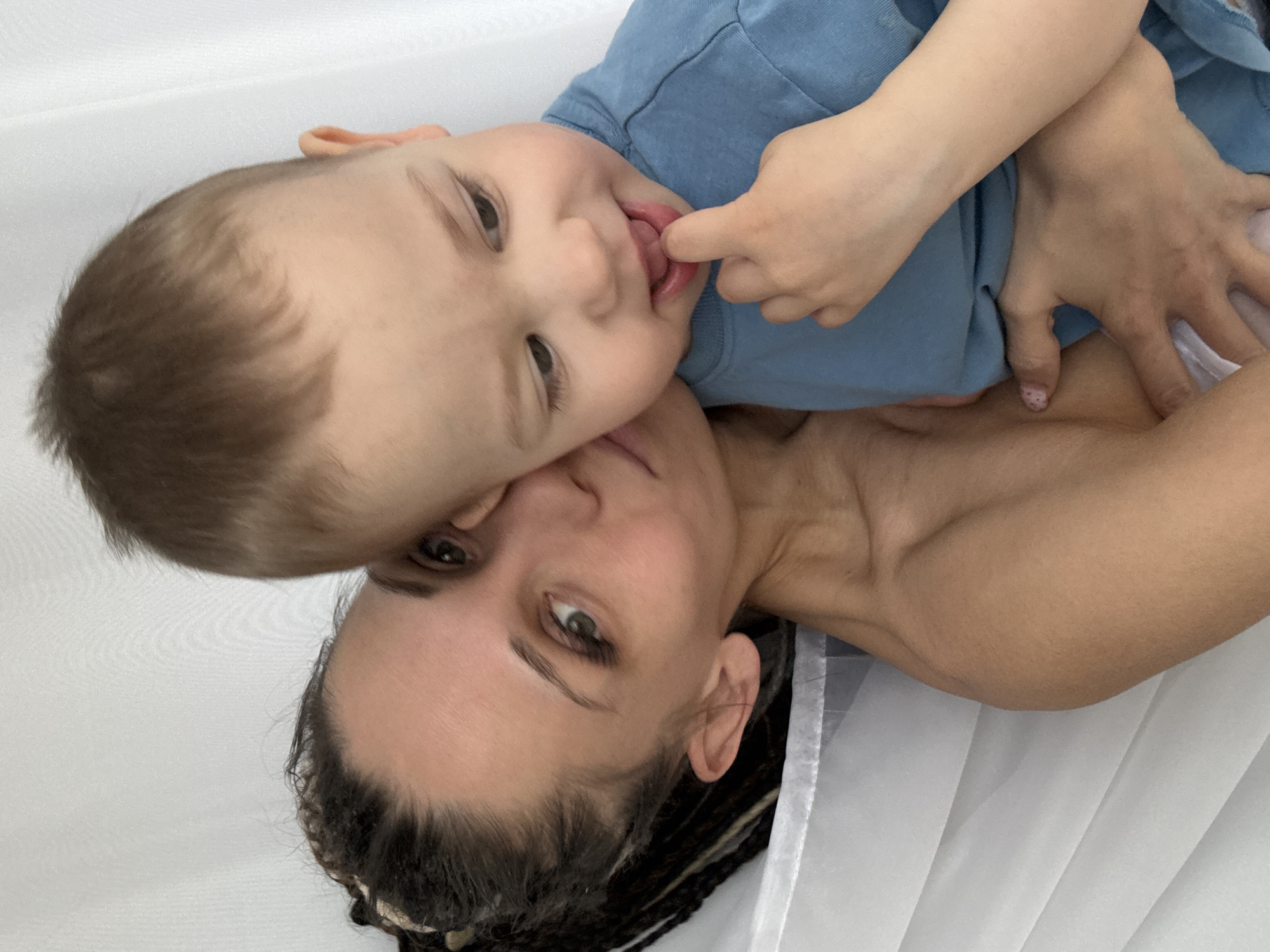 A woman and a young child are lying down. The woman has dark hair in braids and is lying on her back, looking up at the camera. The child is on top of her, wearing a blue shirt, with their face close to the woman's face. The child is sticking their tongue out and touching it with a finger. They are lying on a white surface, possibly a bed or soft pad. The location is not identifiable as Rusanovo, Russia from the given image.