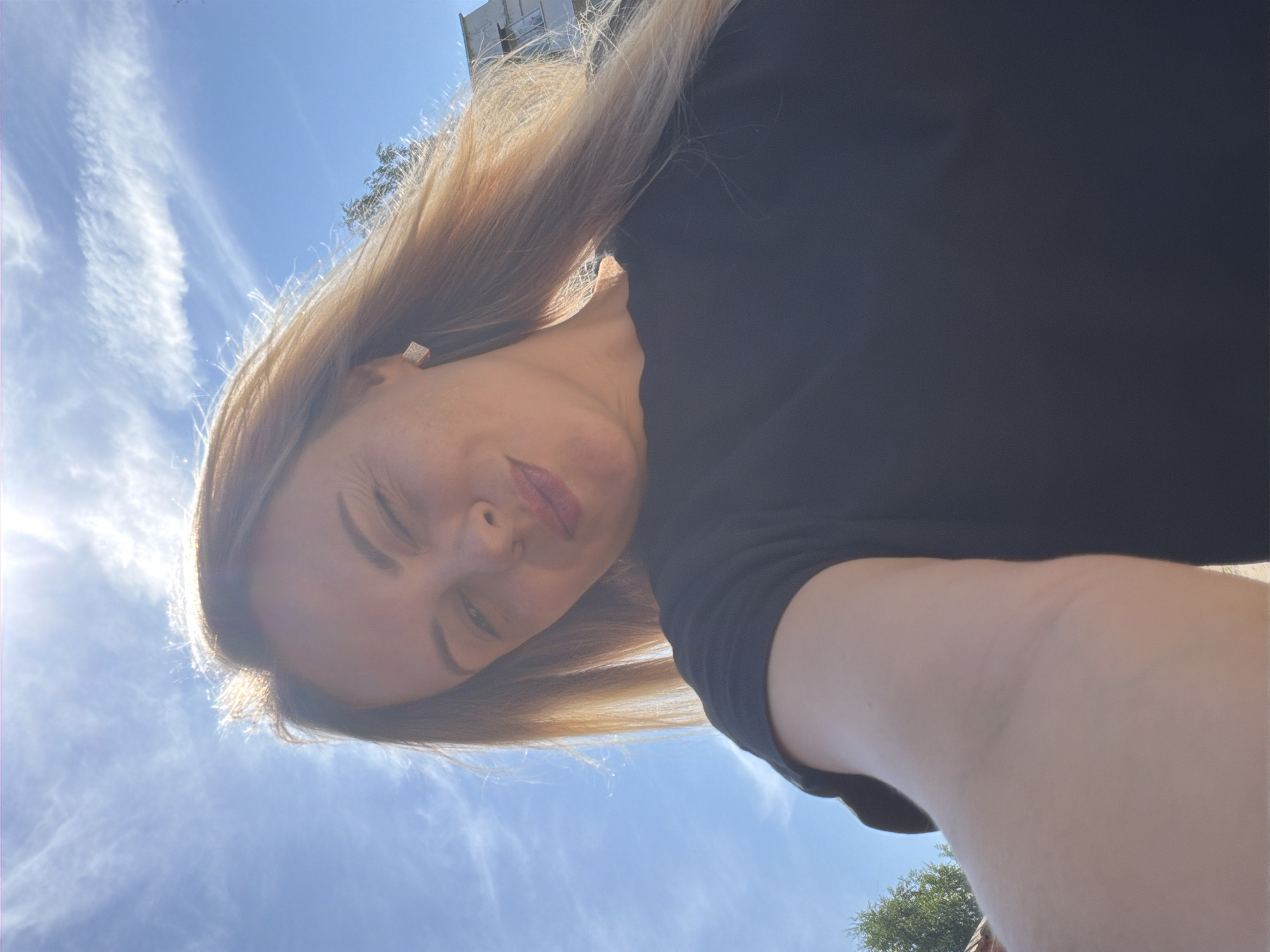 A woman with blonde hair is taking a selfie outdoors under a bright blue sky with wispy clouds. The sun is shining intensely, creating a lens flare effect and casting a warm glow on her hair. She is wearing a black t-shirt and a small square earring. Her right eye is partially closed in a wink, and her lips are slightly pursed. The background shows hints of greenery and a building. The scene appears to be a casual outdoor setting. There is no specific information indicating the location in Votkinsk, Russia.