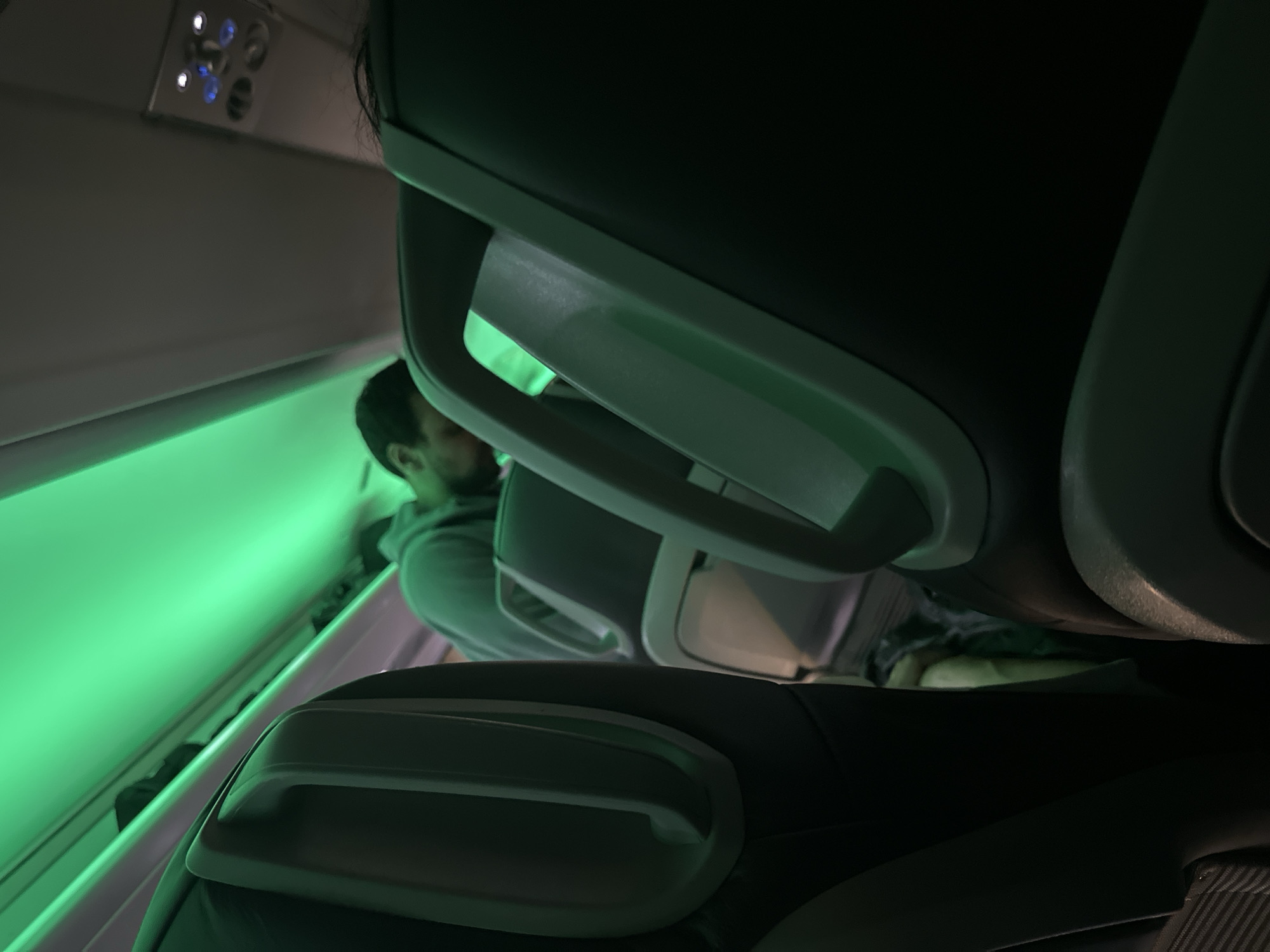The image appears to be taken inside an aircraft cabin, focusing on the back of an airplane seat. The seat is upholstered with a dark material. Visible through the gap between the seat and the headrest of the seat in front is a passenger wearing a gray sweatshirt, looking downward. The cabin lighting emits a green hue. The image is taken from a low angle, suggesting the photographer is seated. The overhead panel is visible in the upper left corner, indicating an airplane setting. There is no evidence to confirm that the location is in the city of Varna, Bulgaria.