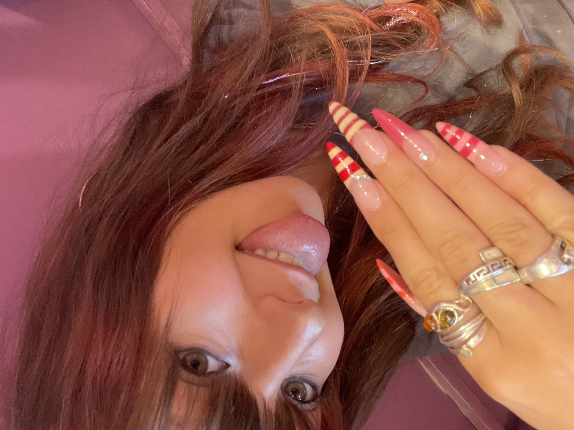 The image shows a close-up of a person's face and hand. The person has brown hair with pink streaks and is wearing a grey shirt. Their right hand, adorned with multiple rings and long, decorated nails, is held up near their face. The nails feature a French tip design with alternating red and white stripes and some with pink tips and small rhinestones. The person's face is tilted, with their eyes open and their tongue sticking out slightly. The background appears to be a pink wall, possibly indoors. There is no indication of the city of Raanana, Israel in the image.