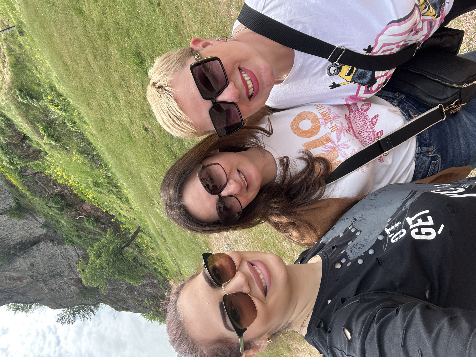 Three women are posing for a selfie. The women are standing in front of the Belogradchik Rocks, a natural rock formation near Belogradchik, Bulgaria. The background includes grass, trees, and the rocky landscape. The woman at the top of the frame has blonde hair and sunglasses, wearing a white t-shirt with a black bag. The woman in the middle has brown hair and sunglasses, wearing a white t-shirt. The woman at the bottom has auburn hair and sunglasses, wearing a black ripped t-shirt. All three women are smiling.