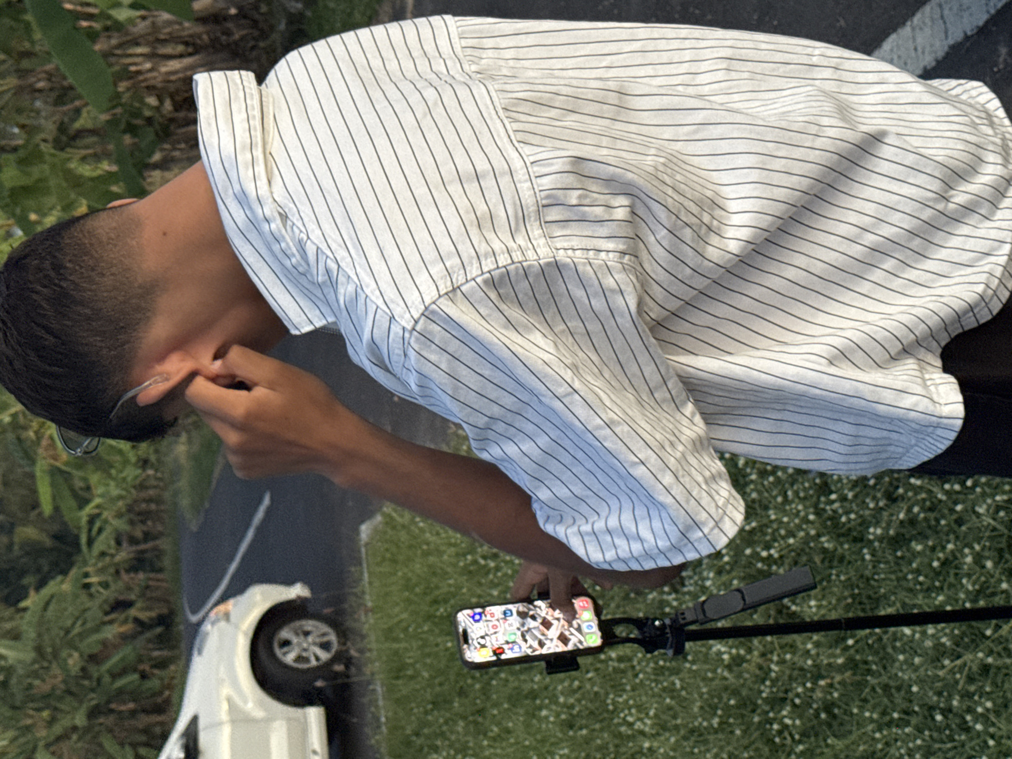 A young man with short dark hair is adjusting his eyeglasses, dressed in a vertically striped white shirt, standing outdoors in Selemadeg Timur, Indonesia. A white car is parked on the asphalt nearby, and a smartphone is mounted on a stand in front of him, on a patch of grassy land.