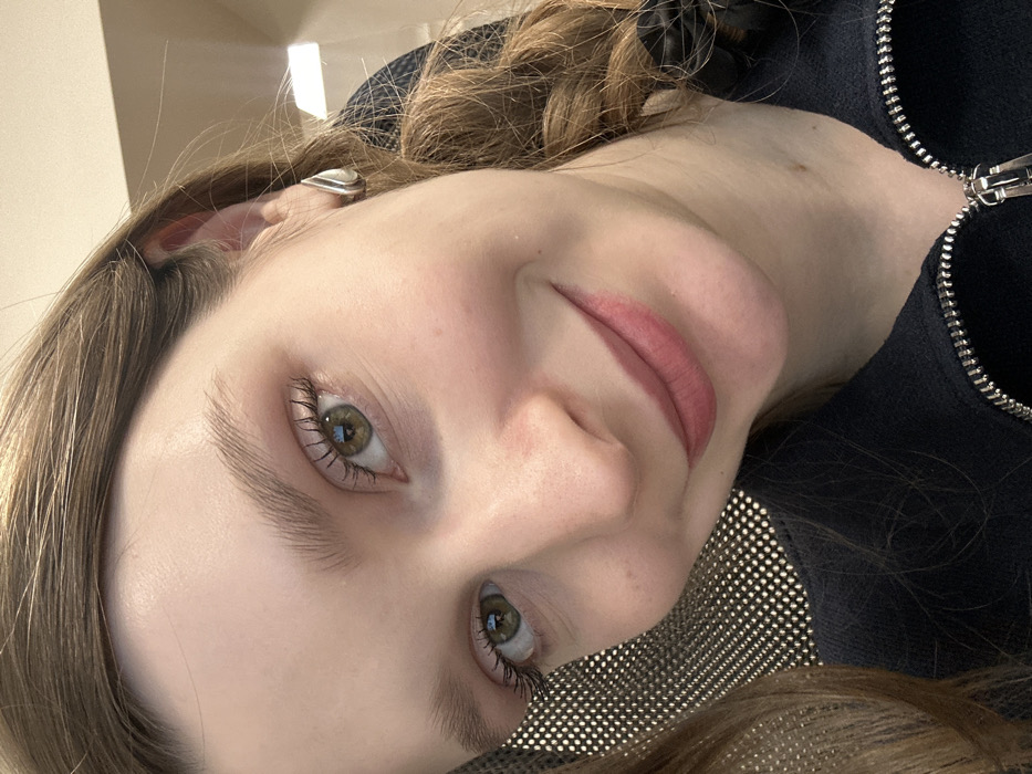 A close-up, eye-level selfie depicts a young Caucasian female in an indoor setting. She has light skin, green eyes with visible eyelashes, and light brown hair partially visible on both sides and braided on the right. She wears a slight, closed-lip smile with light pink lipstick. A silver, teardrop-shaped stud earring is visible on her right earlobe. She is wearing a dark, textured garment, likely a jacket, with a silver zipper partially visible at the lower center. The background is out of focus, showing a grid-patterned surface on the left and a light-colored wall or surface on the upper right. The specific location within the city of Kazan, Russia, is not discernible from the visual information provided.