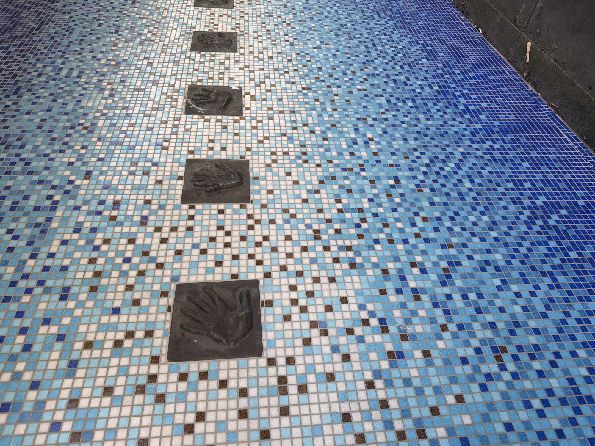 The image captures a section of paved ground, likely a public walkway in Folkestone and Hythe District, United Kingdom. The pavement is covered in a mosaic pattern of small, square tiles in shades of blue, white, and brown. Embedded within the mosaic are a series of square, dark-colored plaques featuring raised impressions of human hands. These plaques are arranged in a linear formation, suggesting a pathway or designated route. The mosaic pattern transitions from a lighter, more varied color scheme in the foreground to a darker, primarily blue hue in the background, potentially indicating a gradient design. The edge of the paved area is visible at the top right corner of the frame, revealing a darker, concrete surface. The overall scene presents a blend of artistic design and practical functionality.