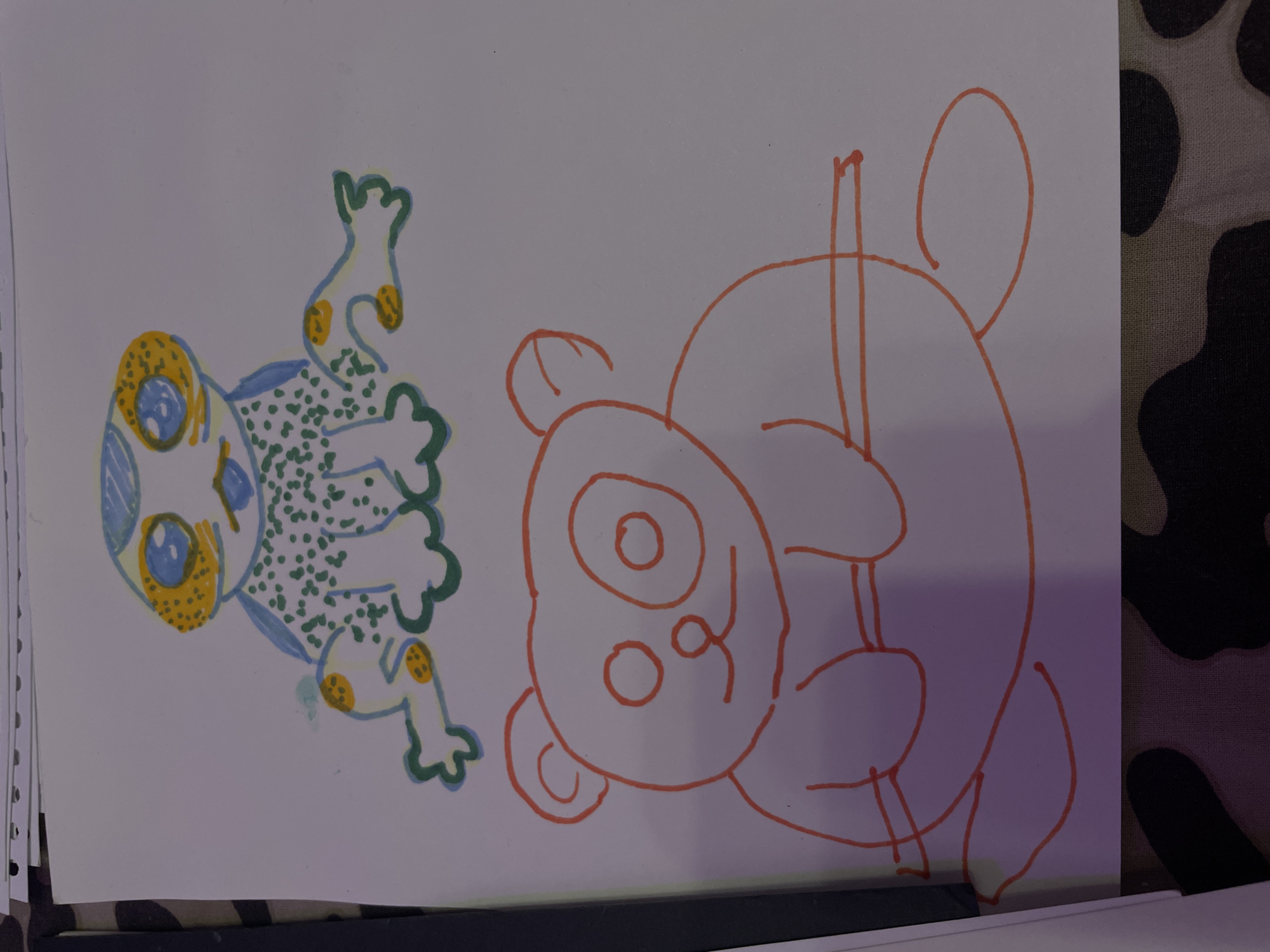 An overhead, slightly angled view captures a child's drawing on a white piece of paper. The drawing features two distinct figures. The upper figure, a frog, is colored with blue for its head, green dots for its body, and yellow and orange accents on its eyes and limbs. The lower figure, an orange outline of a panda, sits with its arms resting on a horizontal line, possibly representing a branch. The paper is resting on a surface with a camouflage pattern. The image does not contain any recognizable landmarks or elements that would allow for identification of a specific city or location like Irinovka, Russia.