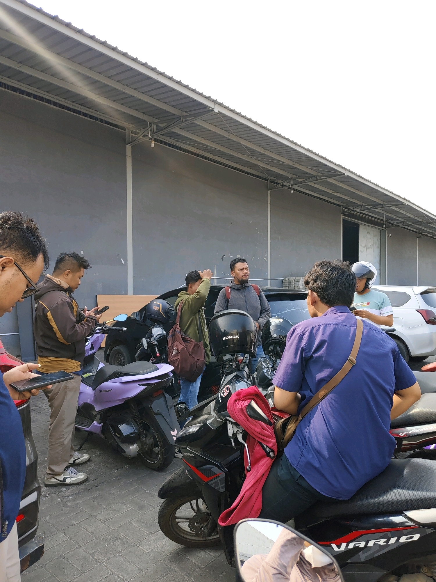 A group of men stand around several parked motorcycles and cars in what appears to be a storage or industrial area, possibly in Probolinggo, Indonesia. A large, grey building with a corrugated metal roof spans the background. In the foreground, several men are interacting, with some looking at their phones. One man in a blue shirt is seated on a black motorcycle, facing away from the camera. Another man in a brown jacket is standing and looking at his phone. A man in a green jacket is holding a brown bag and appears to be looking at something in front of him. Two motorcycles are prominently visible, one light purple and the other black with red accents. The ground is paved with interlocking bricks. The lighting suggests it is daytime, with bright sunlight.