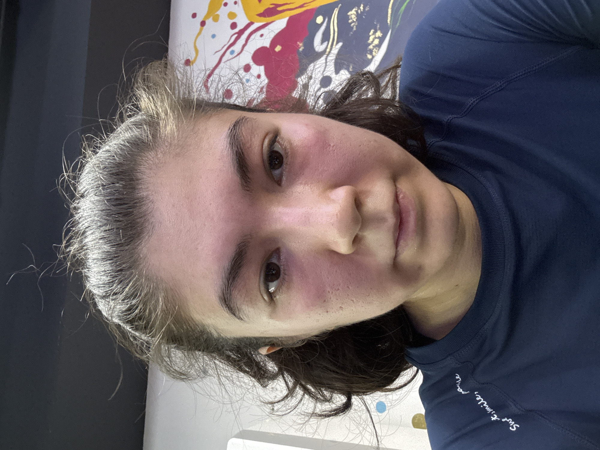 A young woman is shown from the chest up, looking directly at the camera. Her hair is pulled back loosely, with some strands escaping around her face. She has dark eyes, dark eyebrows, and her face appears flushed, particularly on her cheeks. Her skin shows some texture and pores. She is wearing a navy blue long-sleeved shirt. The background is out of focus, but appears to be a wall with abstract artwork featuring splashes of color. The lighting is bright, casting some shadows.