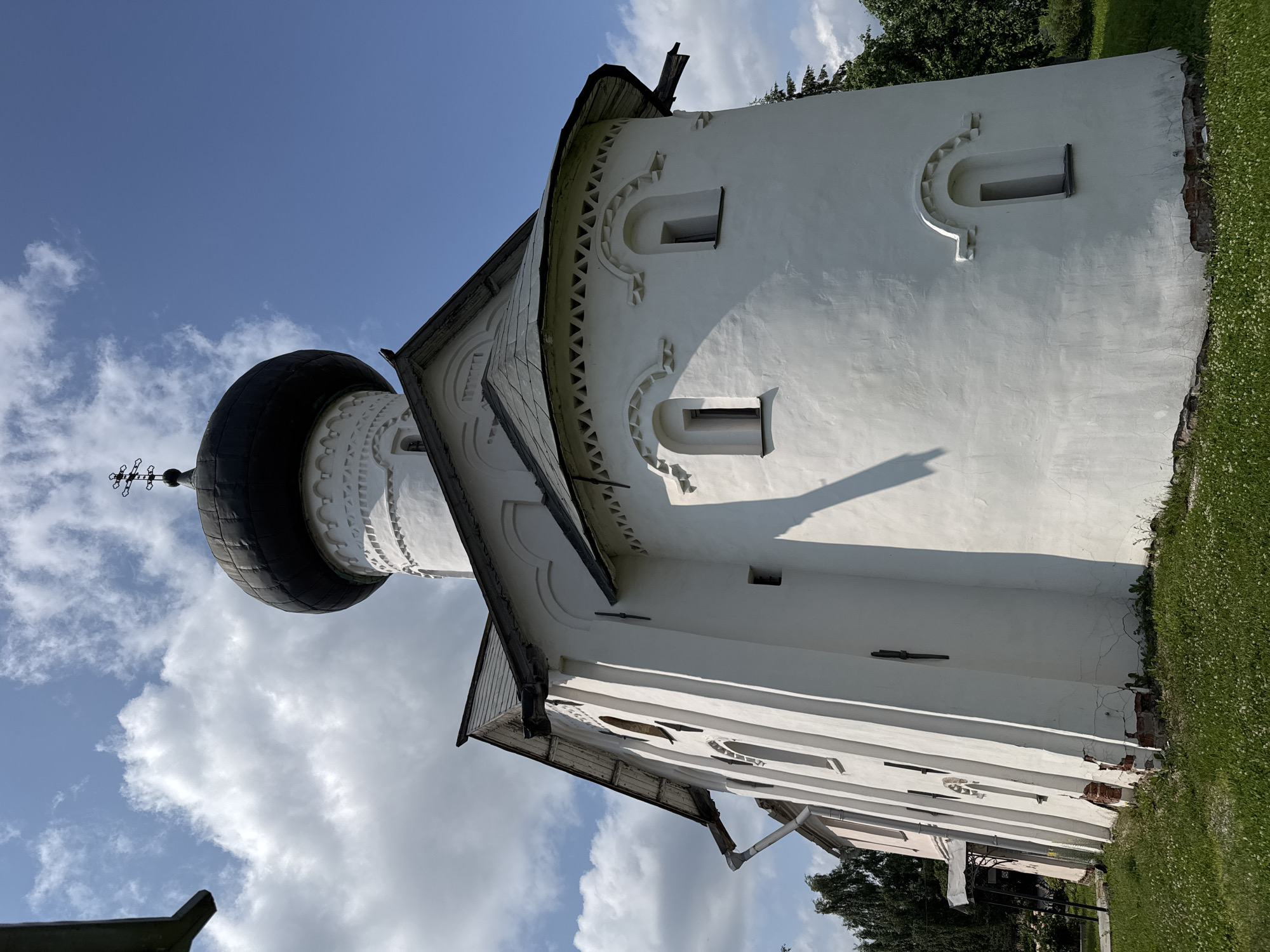 The image shows a white Orthodox church with a dark, onion-shaped dome topped with a cross. The church has a rounded apse with small, arched windows decorated with brickwork patterns. The main body of the church is also white with tall, narrow arched windows. The roof is dark gray. The church is situated on a grassy area with scattered white flowers. In the background, there are green trees and a bright blue sky with white clouds. Part of another building, painted pink, is visible to the left of the church. The architectural style suggests it is a historic church, likely located in Veliky Novgorod, Russia, a city known for its ancient churches. The sunlight casts a shadow on the side of the church. There are no people visible in the image.