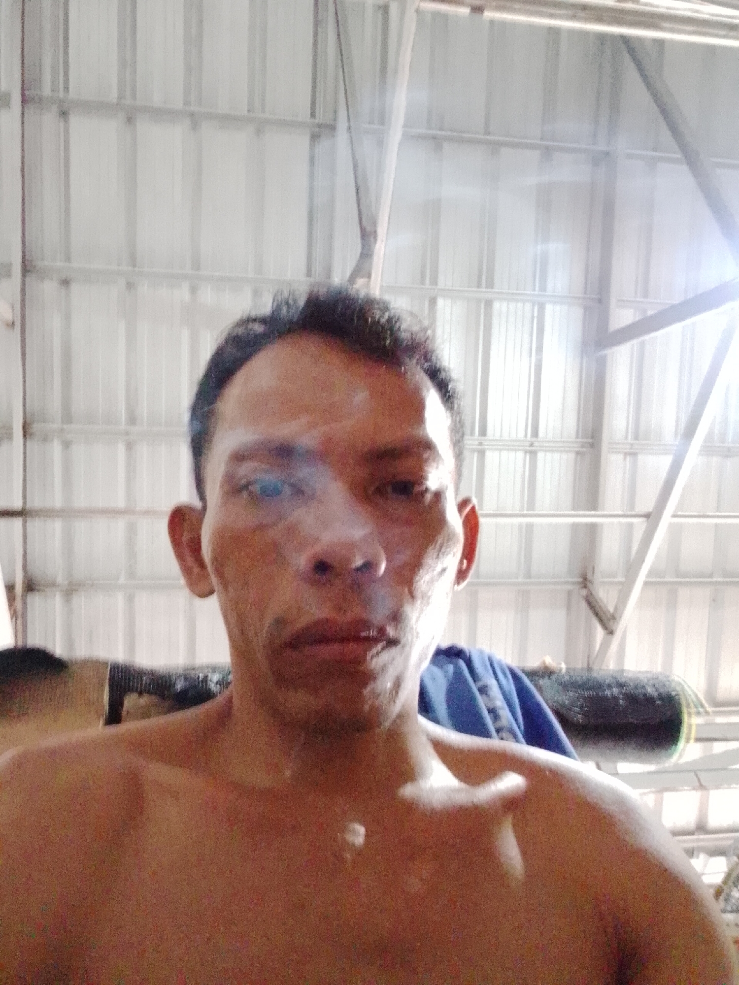A selfie of a middle-aged, dark-haired, bare-chested man against a background of corrugated metal walls and support beams. A blue cloth and a dark, cylindrical object are partially visible behind him. The location may be Bukit Ketok, Indonesia.