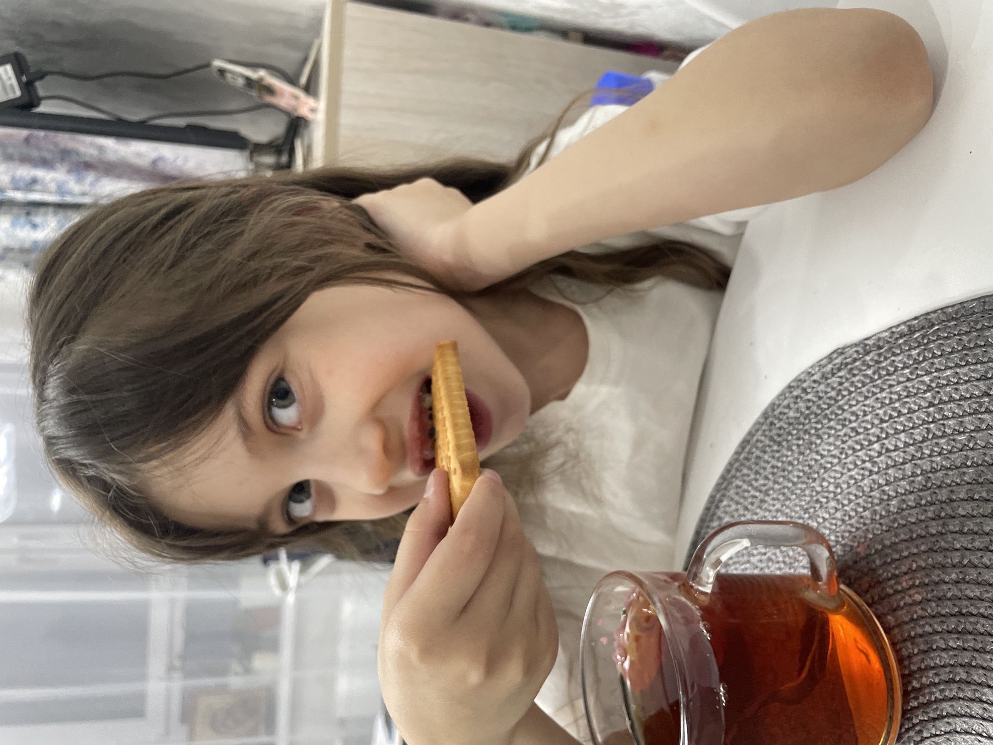A young girl with long brown hair and light-colored eyes is captured indoors, looking directly at the camera with an open-mouthed, engaging expression. She is in the process of eating, holding a rectangular, waffle-patterned cookie (or similar cracker) to her mouth. Her right hand grips the cookie, while her left arm is bent, with her hand near the back of her head. She wears a plain white top.
To the lower right of the frame, a clear glass mug containing a dark reddish-brown liquid, likely tea, sits on a grey, textured placemat. The background is softly blurred but shows elements of an indoor setting, including what appears to be a light-colored shelf or desk with various items like a black electronic device, cables, and other objects, suggesting a casual home environment. The image depicts a candid moment of the girl enjoying a snack and beverage, likely in Izhevsk, Russia. No visible text is present.