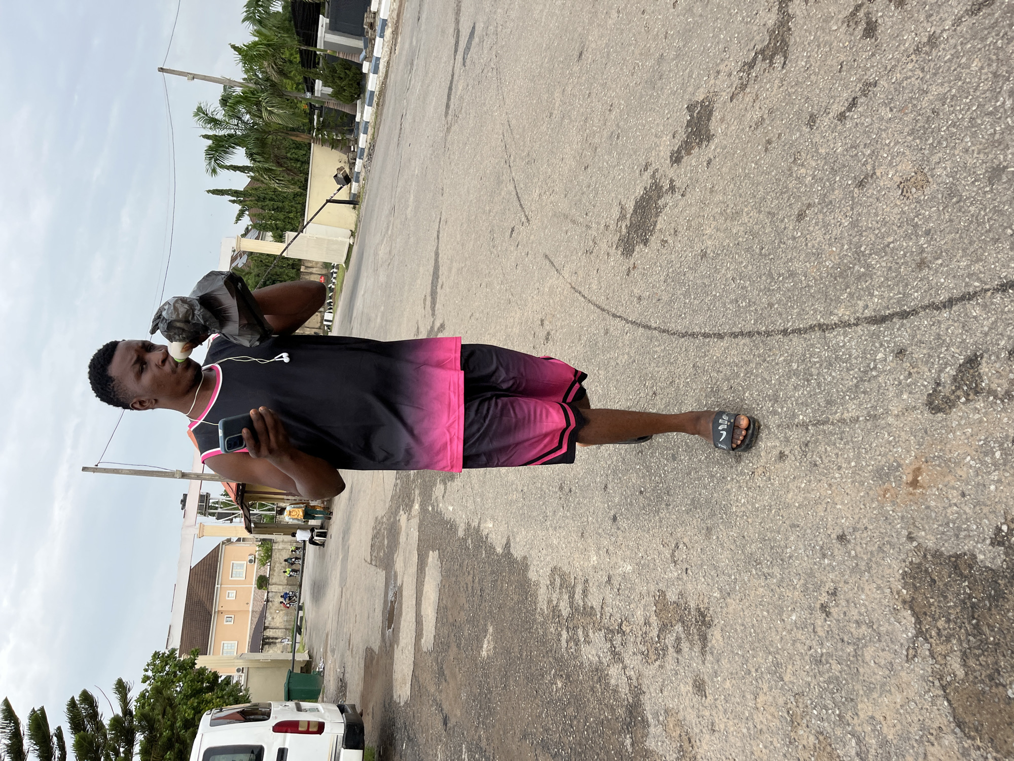 A man stands on an asphalt road. He wears a black sleeveless top with pink trim and matching shorts and is drinking from a plastic bag. He holds a cell phone in his left hand and has earphones in. He wears a sandal on his right foot. The road surface is marked with dark stains and repair patches. In the background are buildings, palm trees, and a white van. This scene is likely in a residential area within the city of Abuja, Nigeria.
