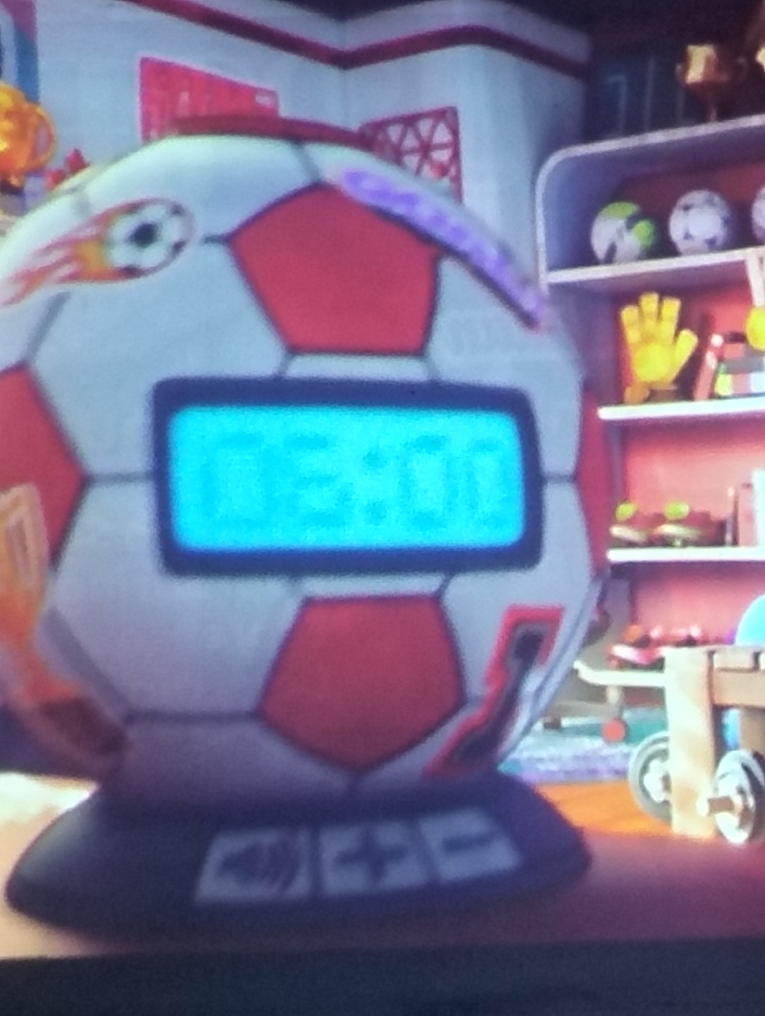 This image captures a close-up of a soccer ball-shaped digital clock. The clock face is illuminated with a bright blue digital display showing the time "03:00". The soccer ball itself is predominantly white with red hexagonal panels and features some colorful graphics, including a soccer ball with flames on one side. The clock sits on a dark base with some white symbols or buttons. In the background, a shelf unit is visible with various items, including what appear to be small soccer balls, a yellow glove, and some trophies or decorative objects. The overall environment suggests a child's room or a playful setting. There is no indication of the city of Payagaboh, Indonesia, within the image.