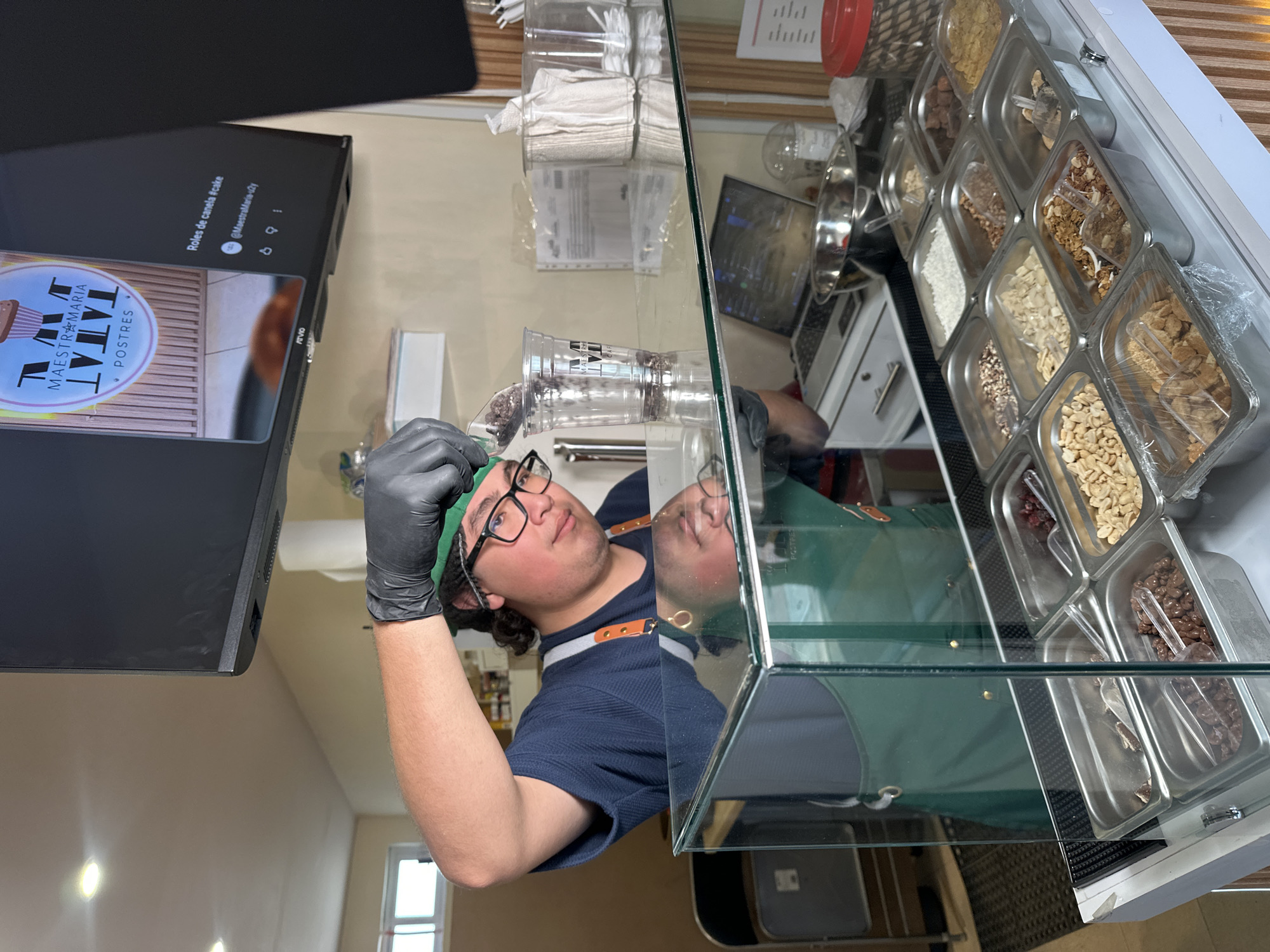 The image shows a person preparing a dessert order in what appears to be a food establishment, potentially located in San Andrés Cholula, Mexico, based on the brand/logo visible. The person is wearing a blue shirt, green apron, black gloves, and glasses, and has a green hair covering. They are holding a clear plastic cup filled with small, dark-colored toppings, likely chocolate chips or similar candies, ready to add to an order.
Behind the person, there's a glass display case filled with various toppings in individual containers, including nuts, coconut flakes, chocolate pieces, and other ingredients used for dessert customization. There's a TV screen on the left side of the image, displaying a logo and social media handle, possibly for the business where the image was taken. The logo is "MAESTRA MARIA POSTRES" (desserts). The social media handle displayed is "@MaestraMaria-uzy," and the text says "Roles de canela #cake" (Cinnamon rolls #cake). The environment suggests a professional food preparation area.