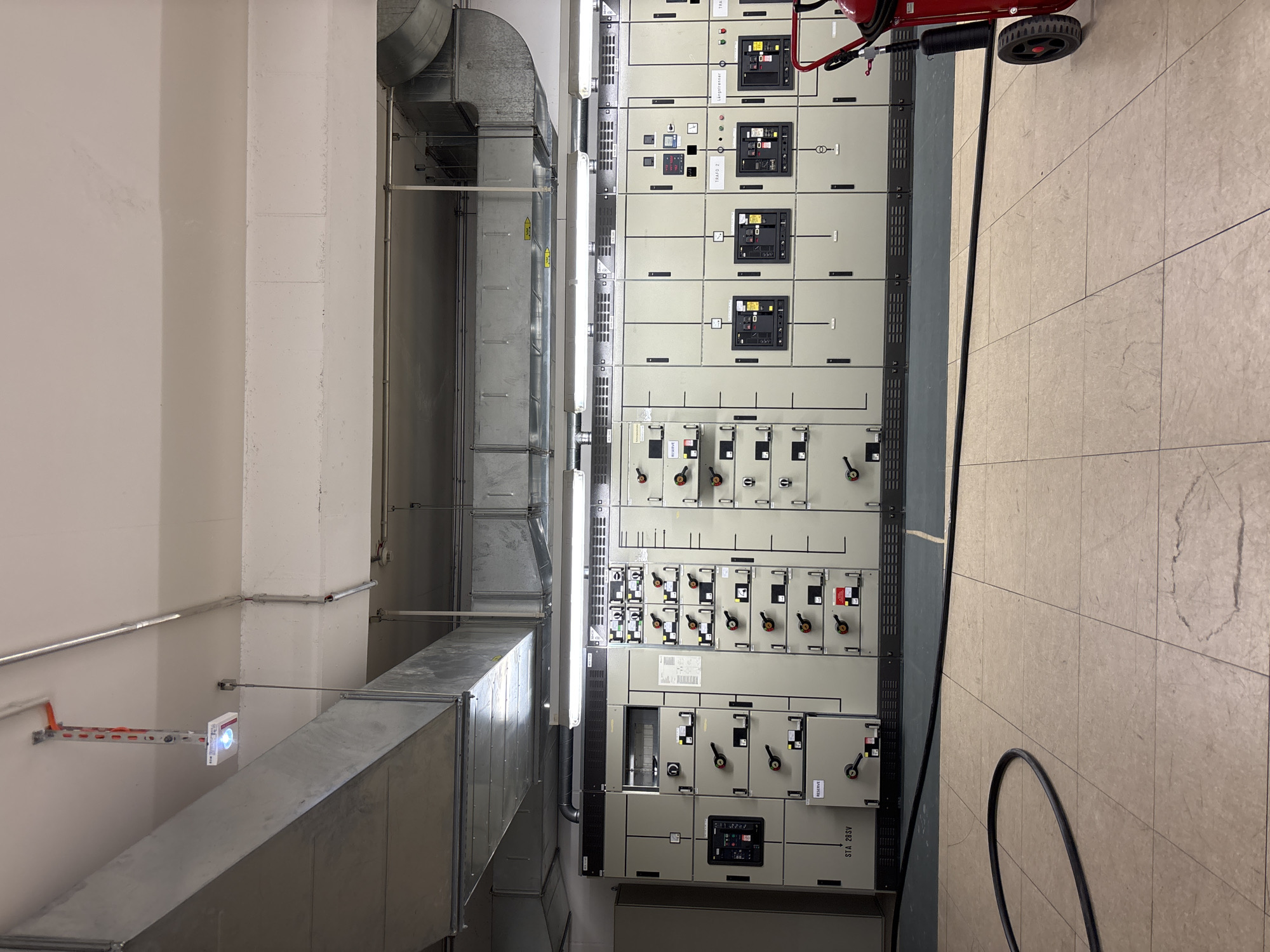This image depicts an interior view of an electrical control room, likely located in a commercial or industrial facility within the city of Mannheim, Germany. Dominating the scene is a large, modular electrical panel system extending horizontally across the frame. The panels are light gray with numerous compartments, each featuring black levers, switches, and indicator lights, suggesting active electrical equipment. Some compartments have labels, though they are largely illegible from this distance. Above the panels, industrial ventilation ducts made of galvanized metal are prominently installed on the ceiling, along with fluorescent lighting fixtures. In the foreground, a black electrical cable is coiled on the tiled floor. To the far right, a red fire extinguisher on wheels is partially visible. The overall environment is utilitarian and functional, characteristic of a power distribution or control center.